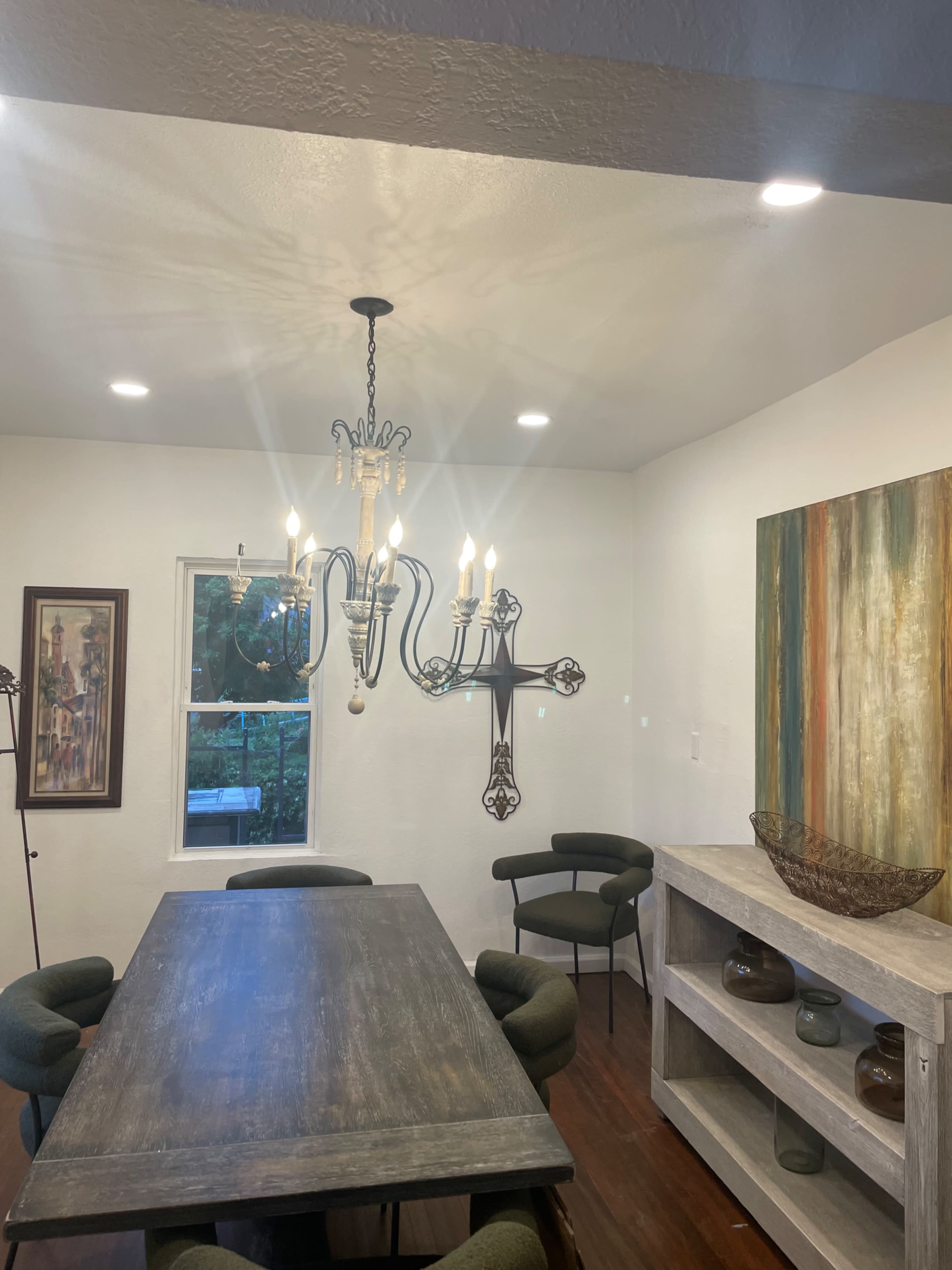 The image shows a dining area with a wooden table, green upholstered chairs, a chandelier, and framed artwork on the walls.