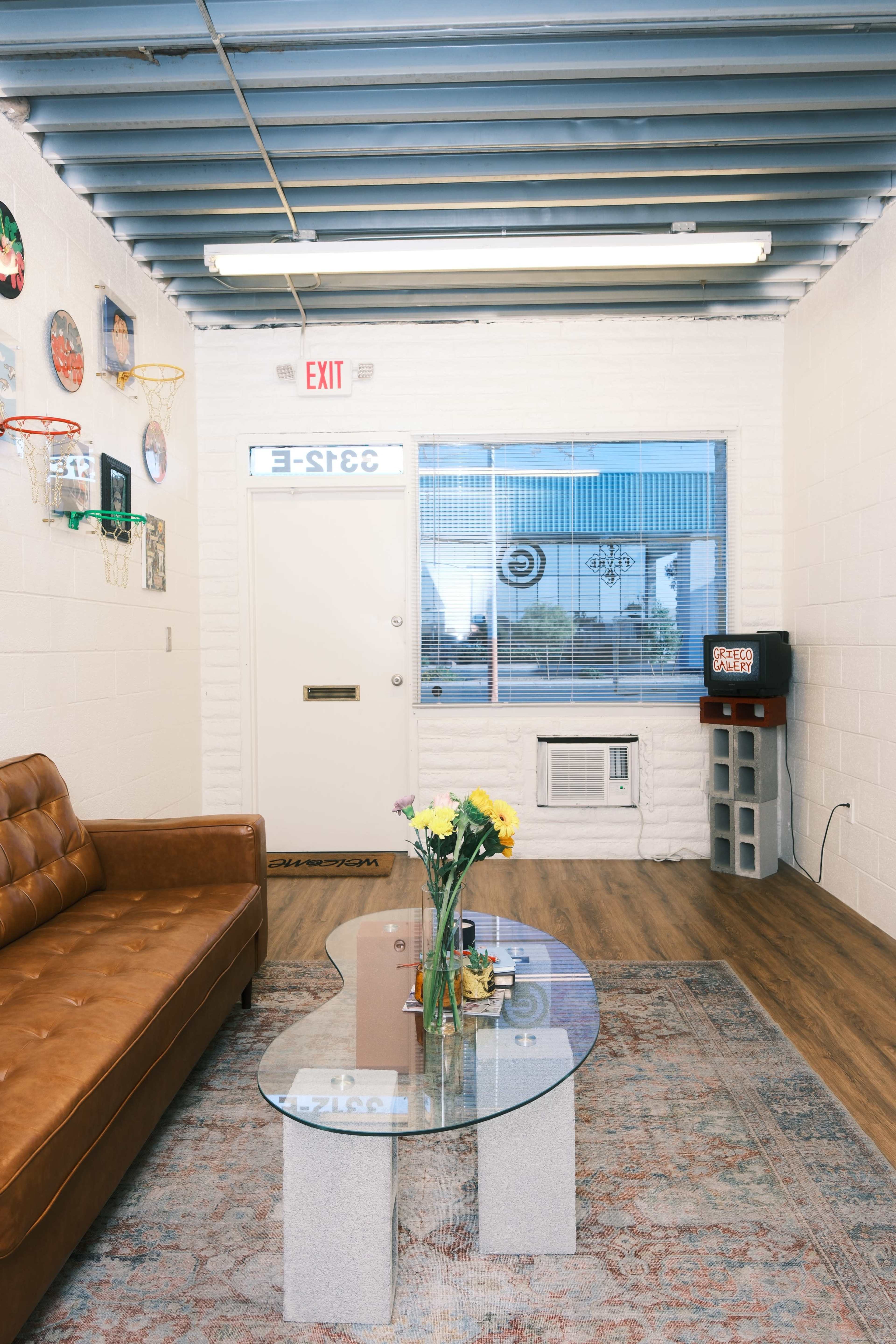 The image shows a small indoor space with a brown leather couch, a glass coffee table, a window, and a wall-mounted air conditioning unit.