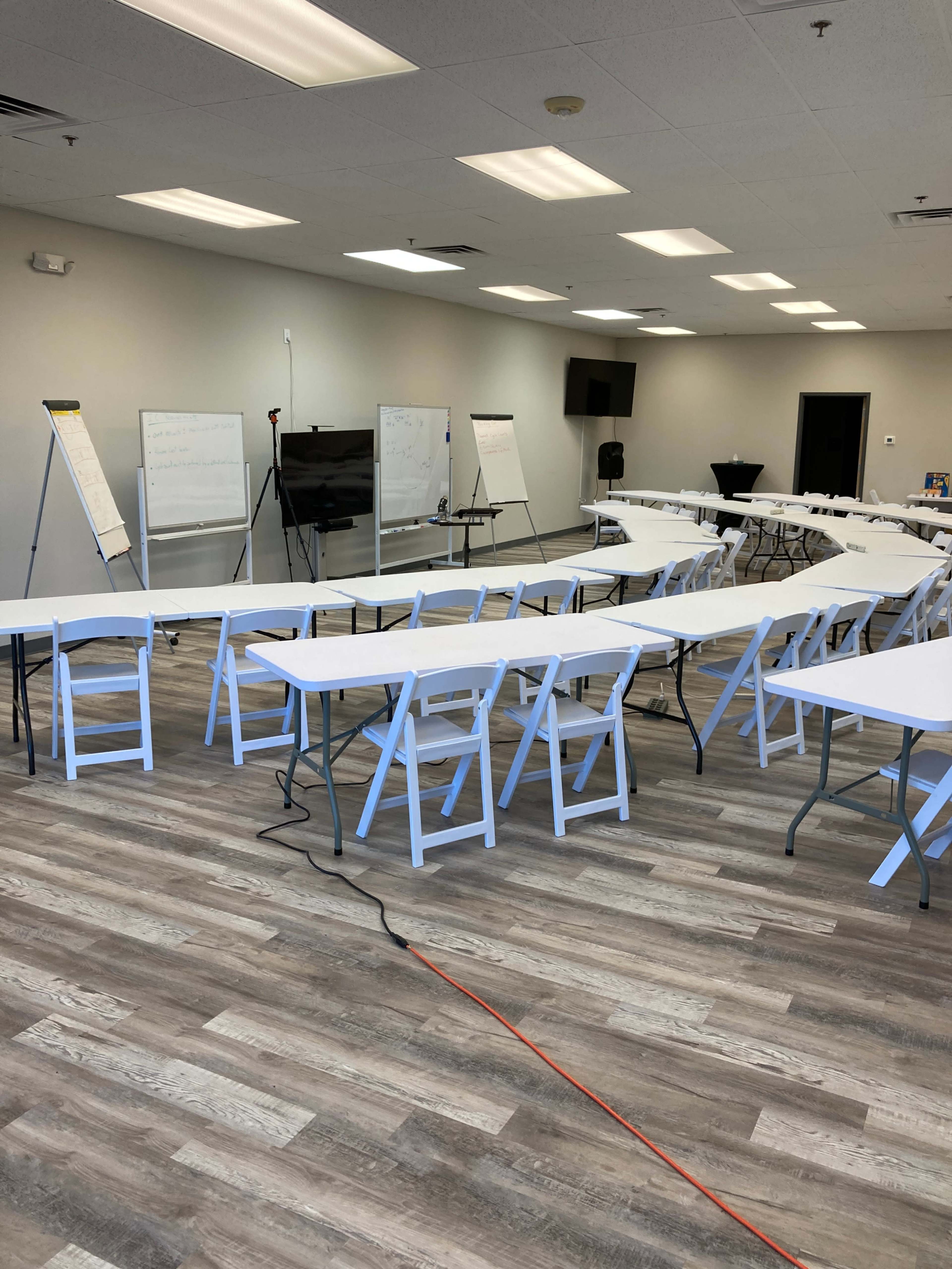 A spacious training room contains several white tables arranged in rows, along with flip charts and a presentation screen at the front.
