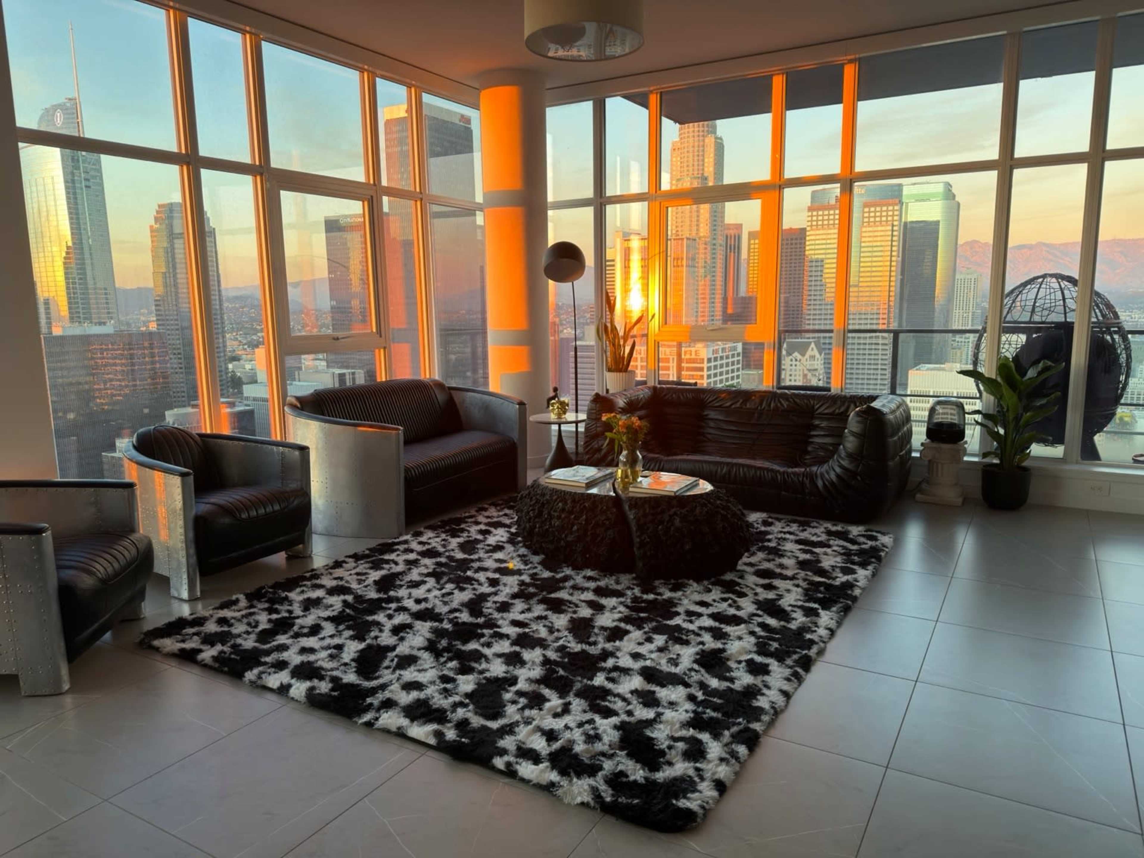 A modern living room features black leather furniture, a circular coffee table, and a large area rug with a cow print, all illuminated by sunset reflections from the city skyline outside.