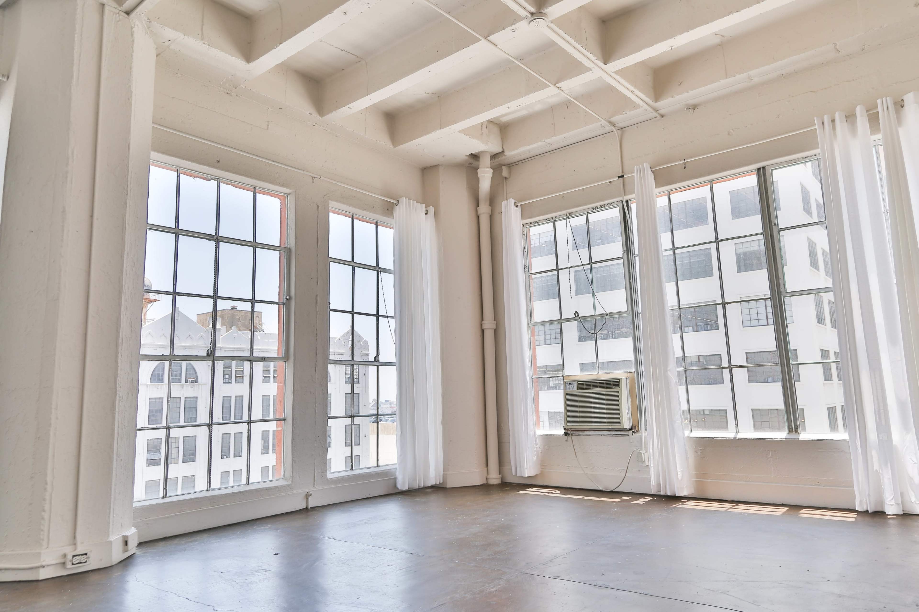 An empty room with large windows featuring sheer white curtains and concrete floors, complemented by an air conditioning unit mounted in one of the windows.