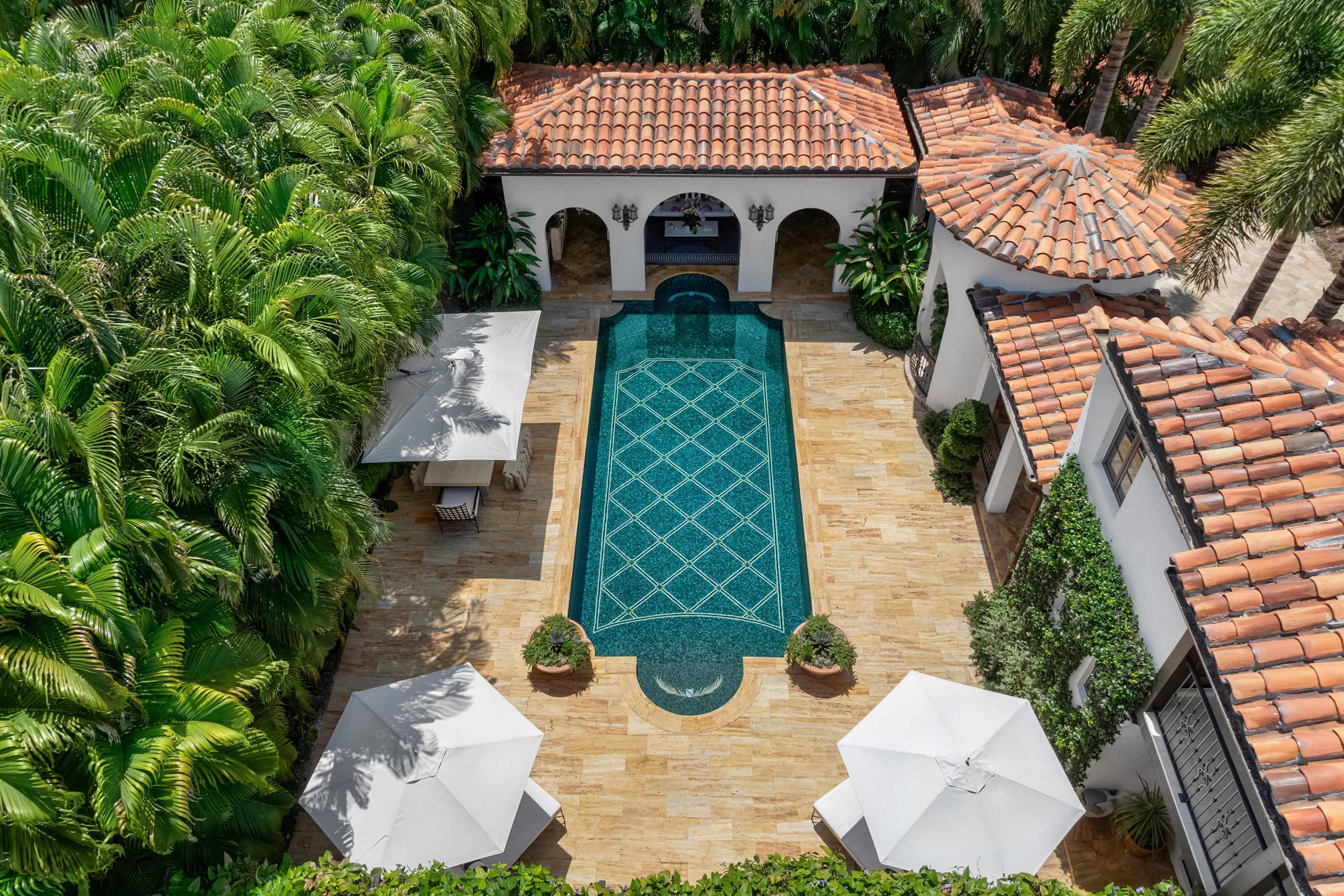 The image shows a rectangular swimming pool surrounded by tropical greenery and stone pathways, with shaded seating areas featuring umbrellas.