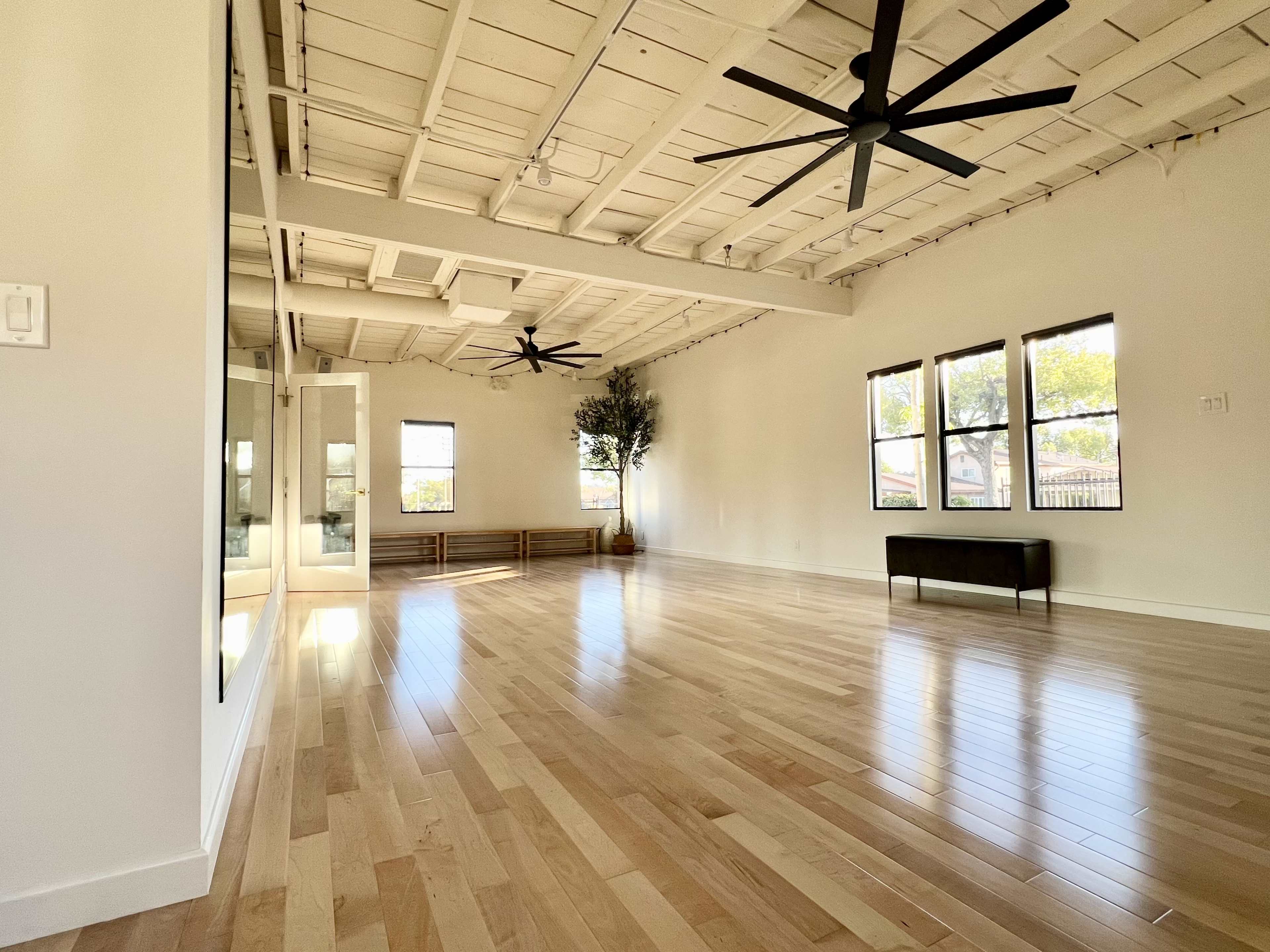 A spacious, well-lit room with hardwood flooring, two ceiling fans, and large windows.