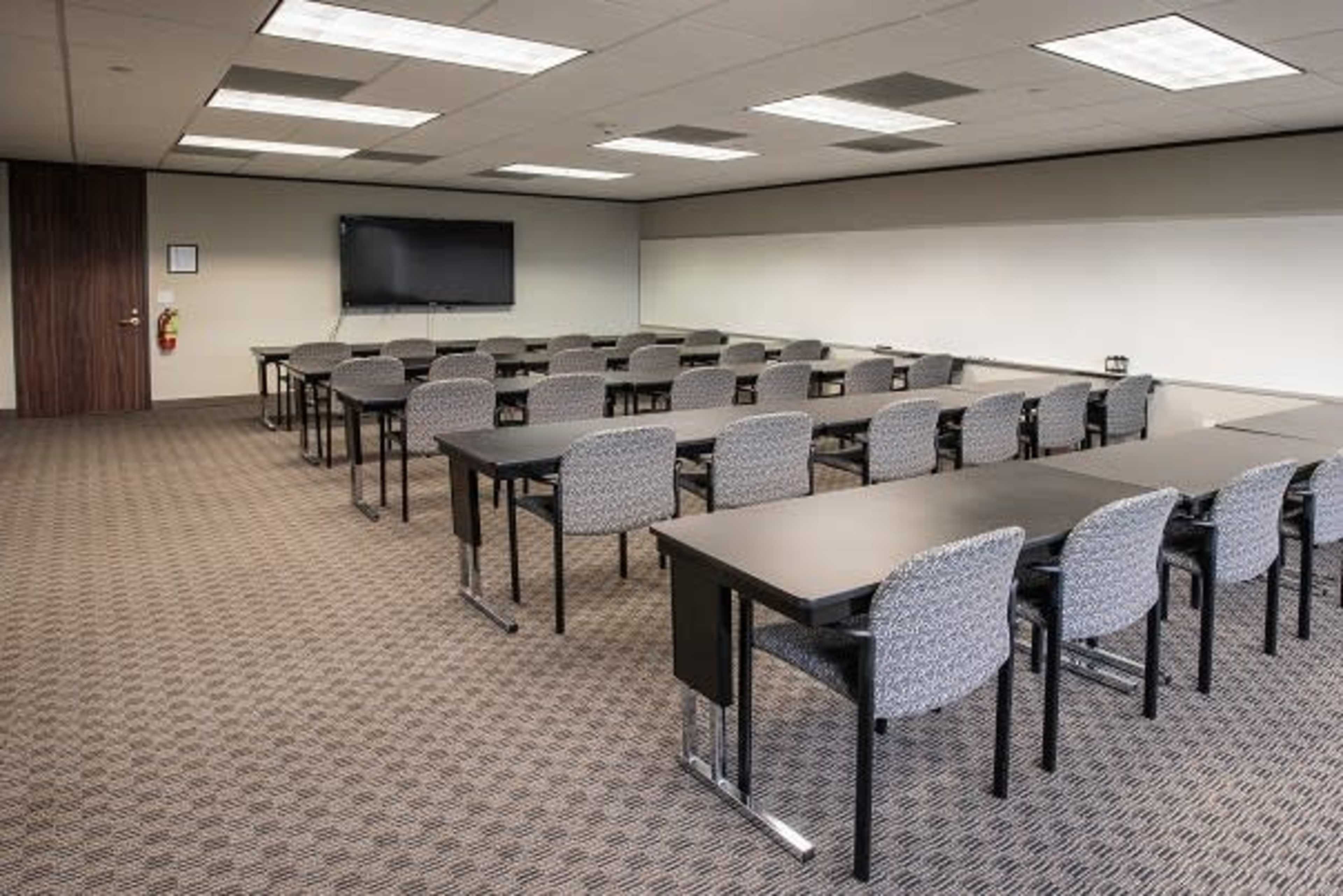 A conference room with multiple rows of tables and chairs facing a wall-mounted television.