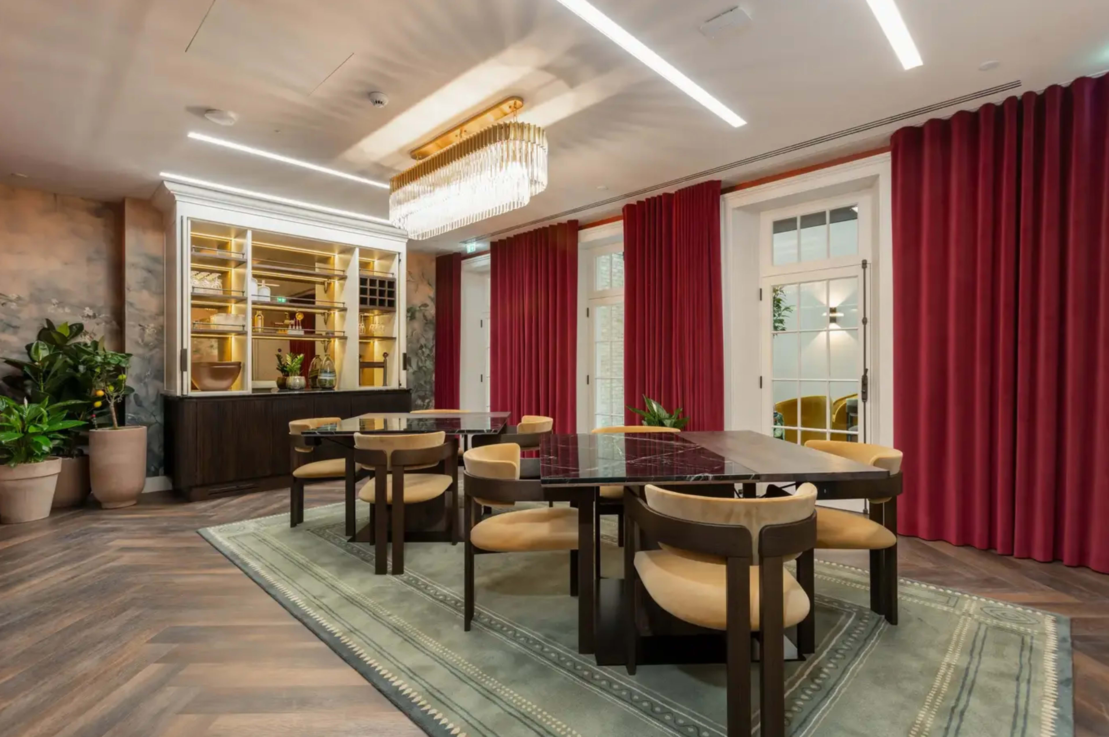 The image shows a dining area with a large rectangular table surrounded by beige chairs, a patterned rug beneath, and a chandelier above, set against red curtains and a cabinet in the background.