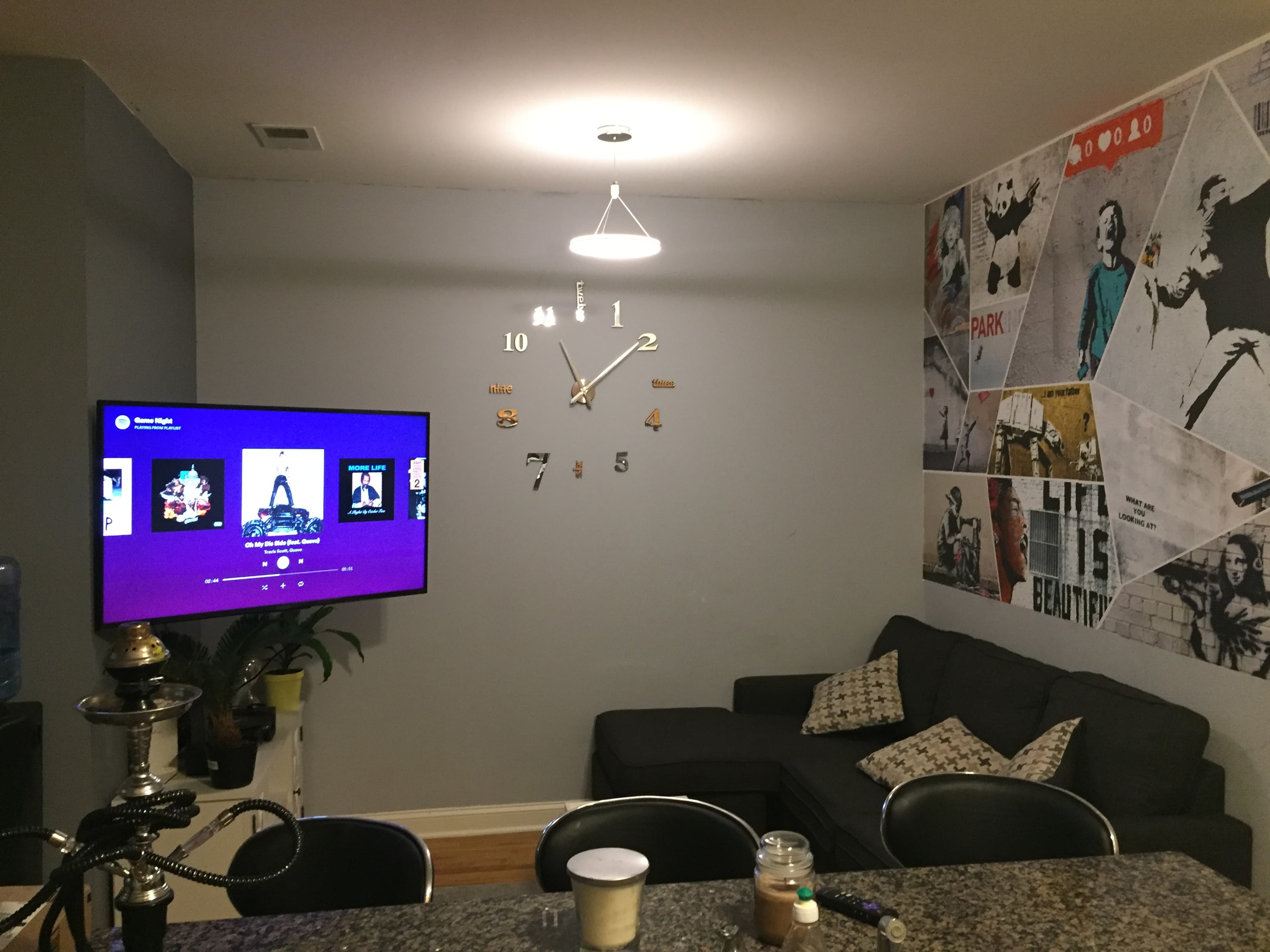 A living room features a modern clock on the wall, a television displaying artwork, and a dark sofa with patterned cushions.