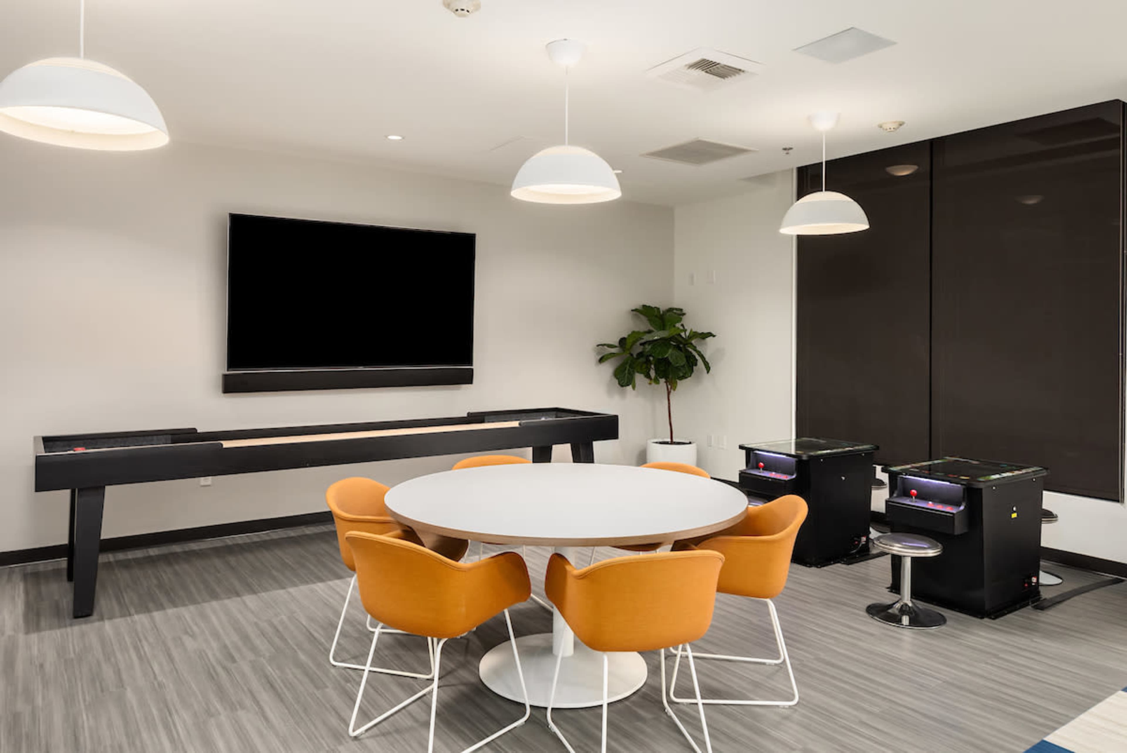 A modern recreation room features a round table with orange chairs, a large TV screen mounted on the wall, and shuffleboard and arcade games against one side.