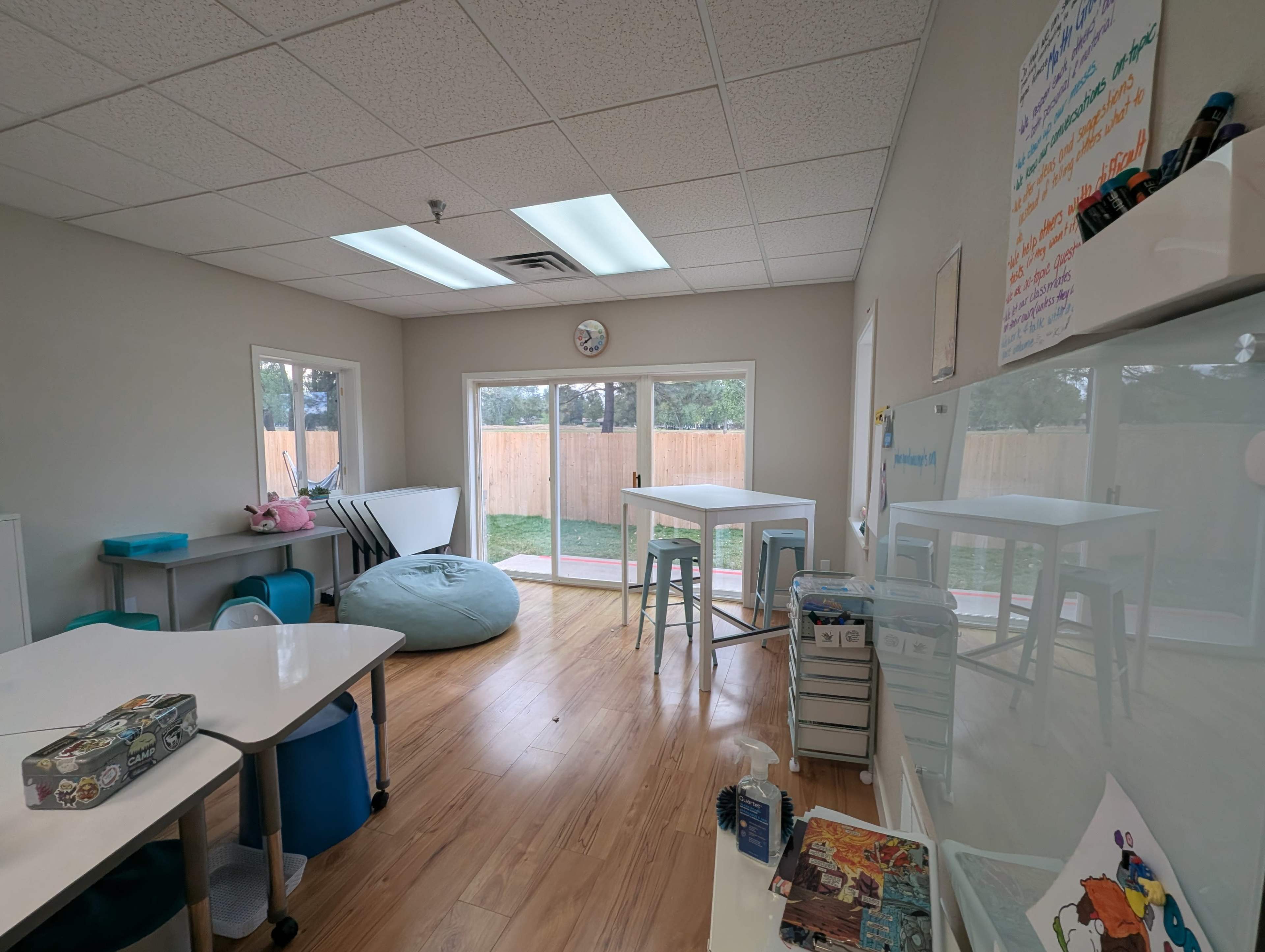 The image shows a bright, uncluttered classroom with tables, a bean bag chair, and large windows overlooking a fenced outdoor area.