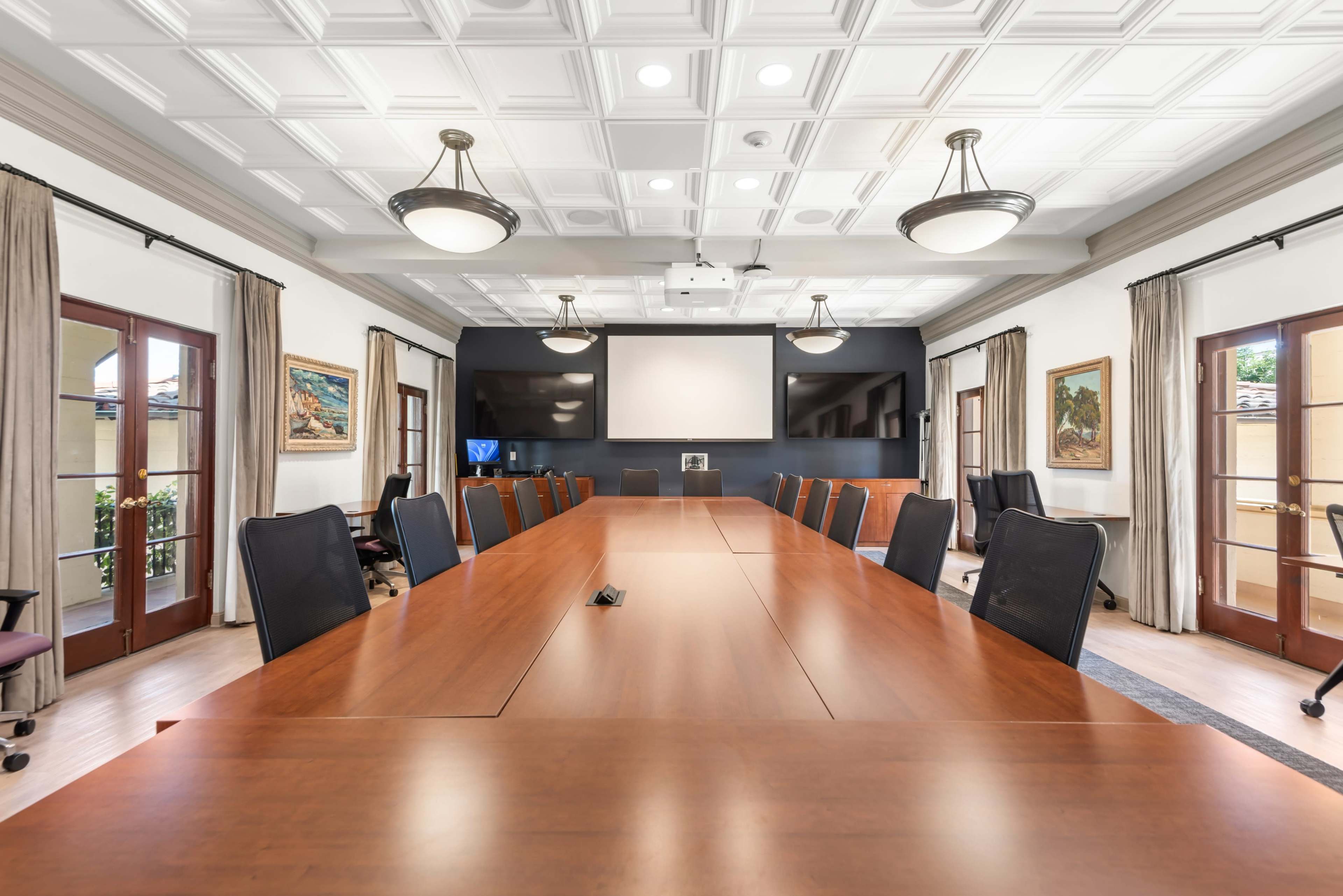 A large conference room features a long wooden table surrounded by black chairs, with a projector screen and doors leading to an outdoor area.