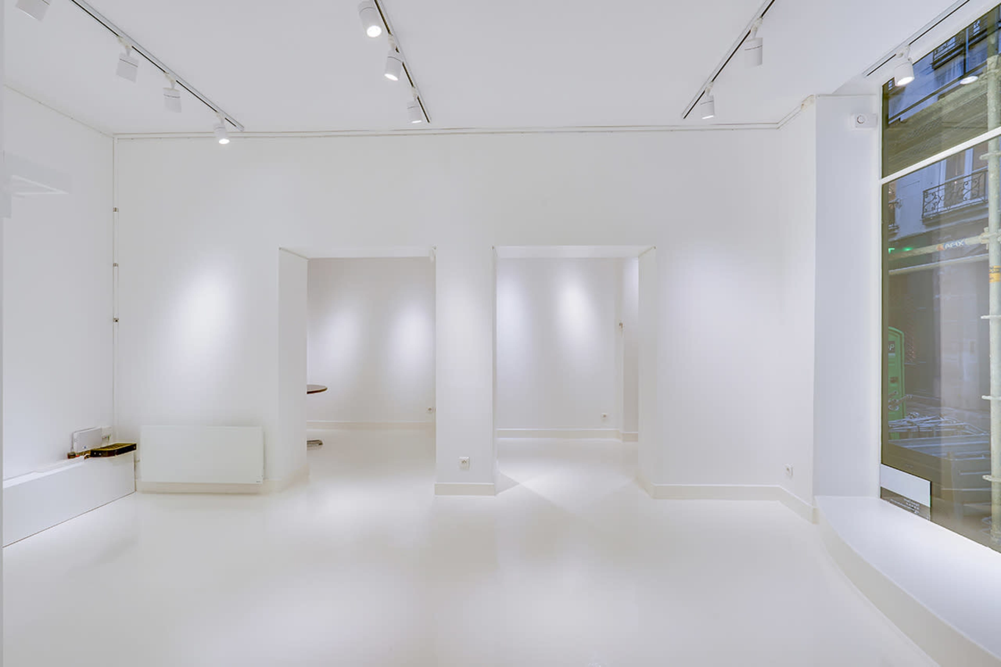 The image shows a spacious, minimalist gallery with white walls and a polished floor, featuring several recessed lighting fixtures and large windows.