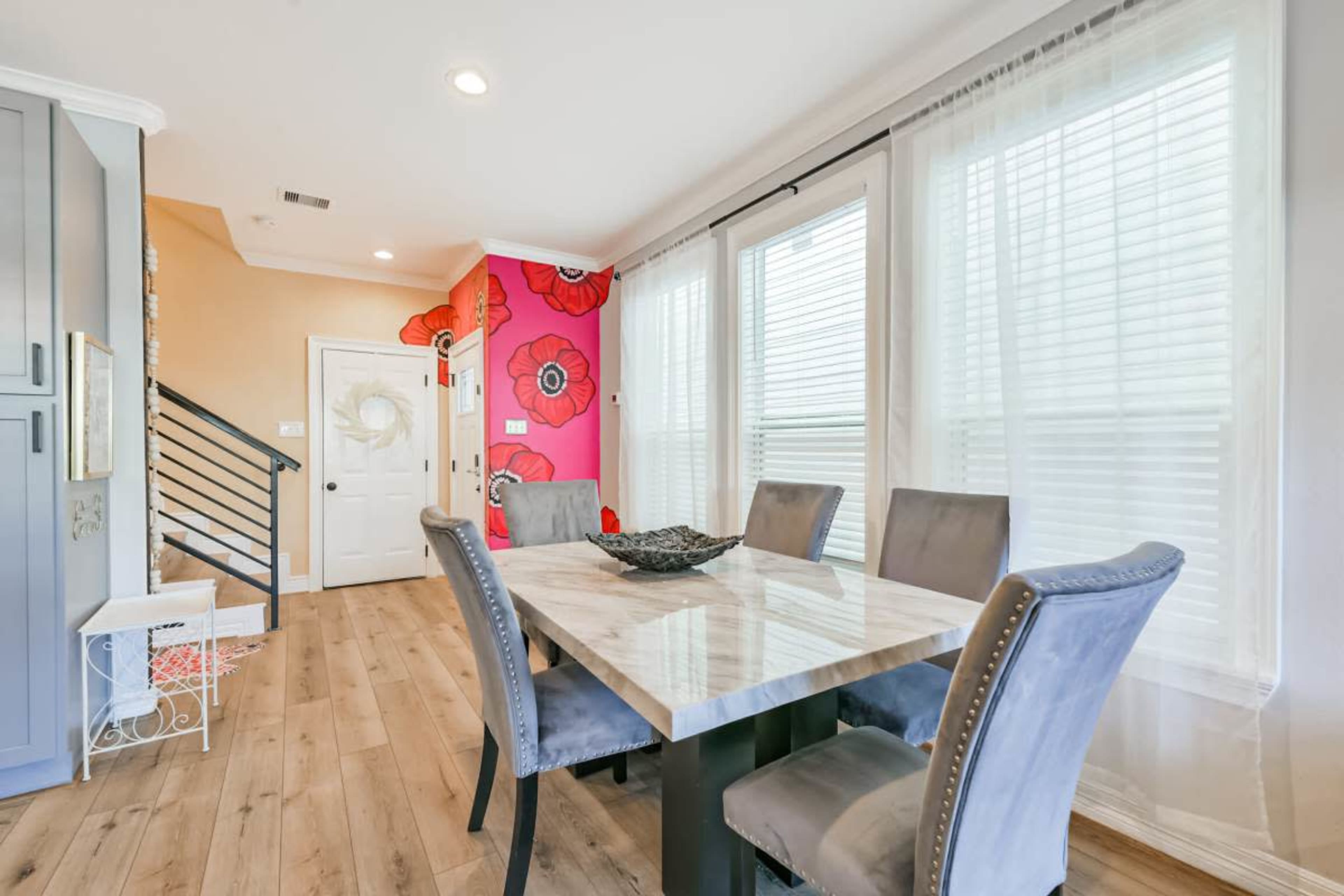 The image shows a dining area featuring a marble top table with gray upholstered chairs, situated next to large windows with sheer curtains and a colorful floral accent wall.