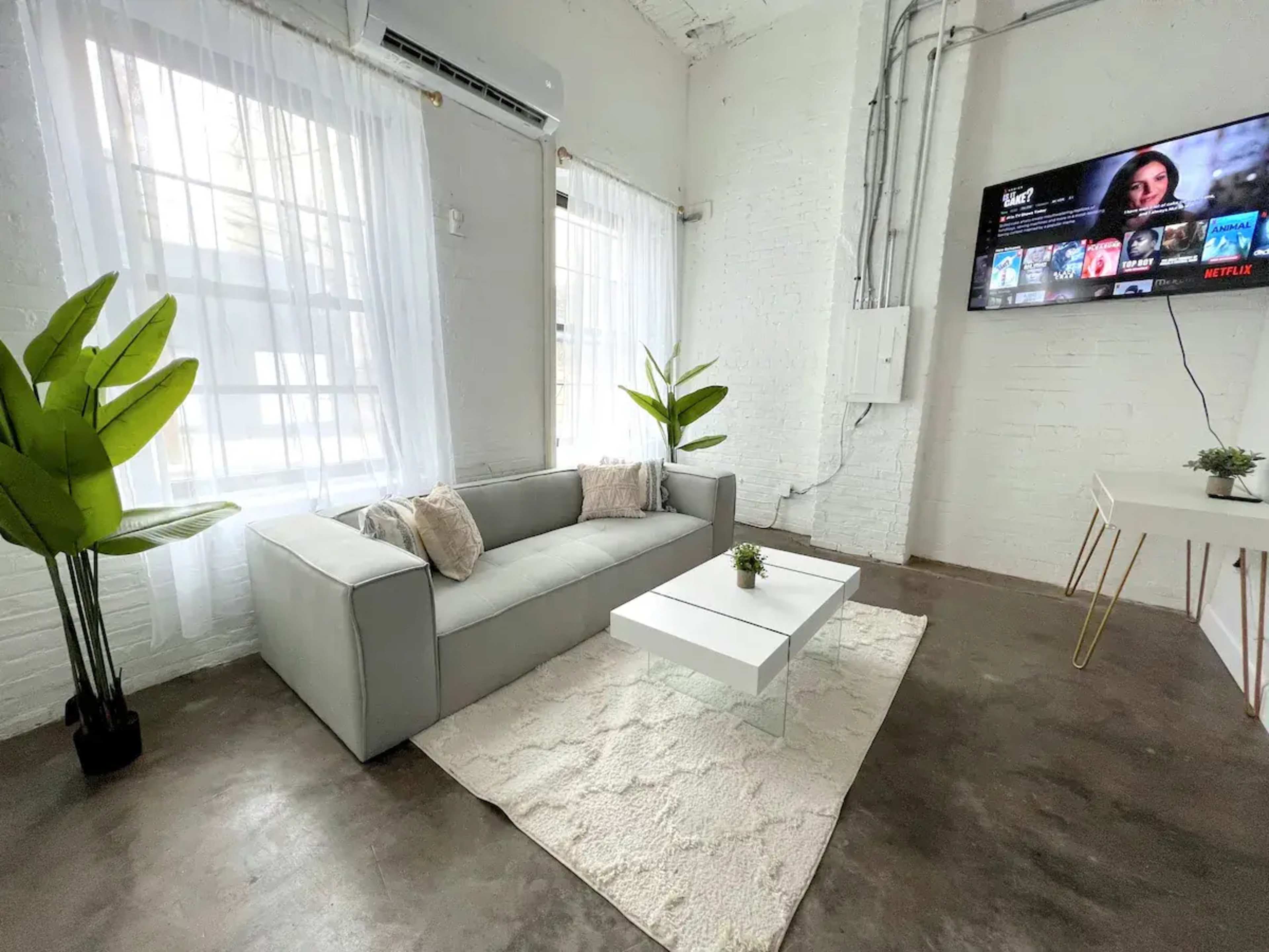 A modern living room with a gray couch, a white coffee table on a rug, and a wall-mounted television displaying a Netflix interface.