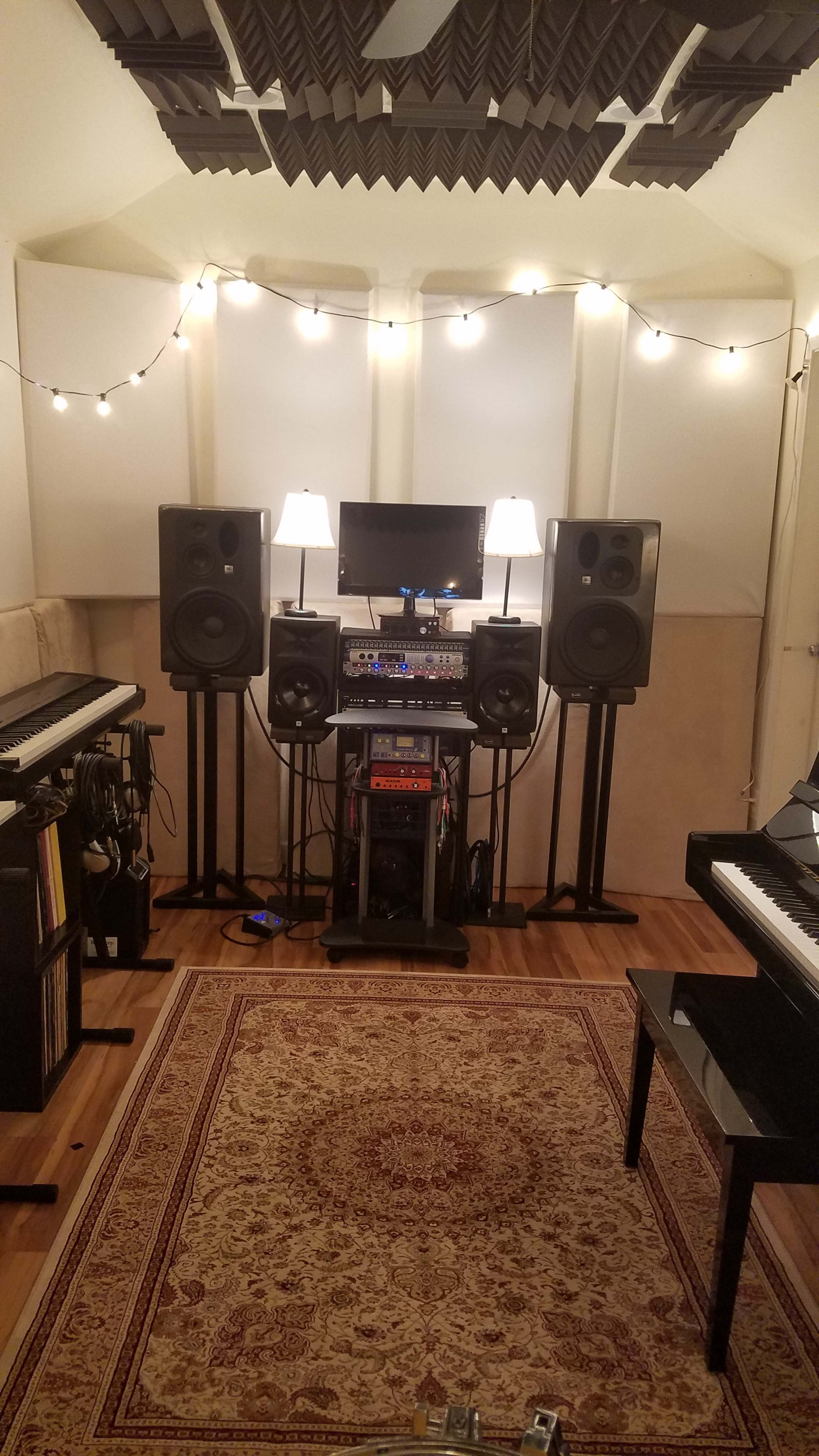 A home recording studio featuring two large speakers on stands, a central mixing console, a monitor, a black piano, and decorative string lights in a cozy room with hardwood flooring and a rug.