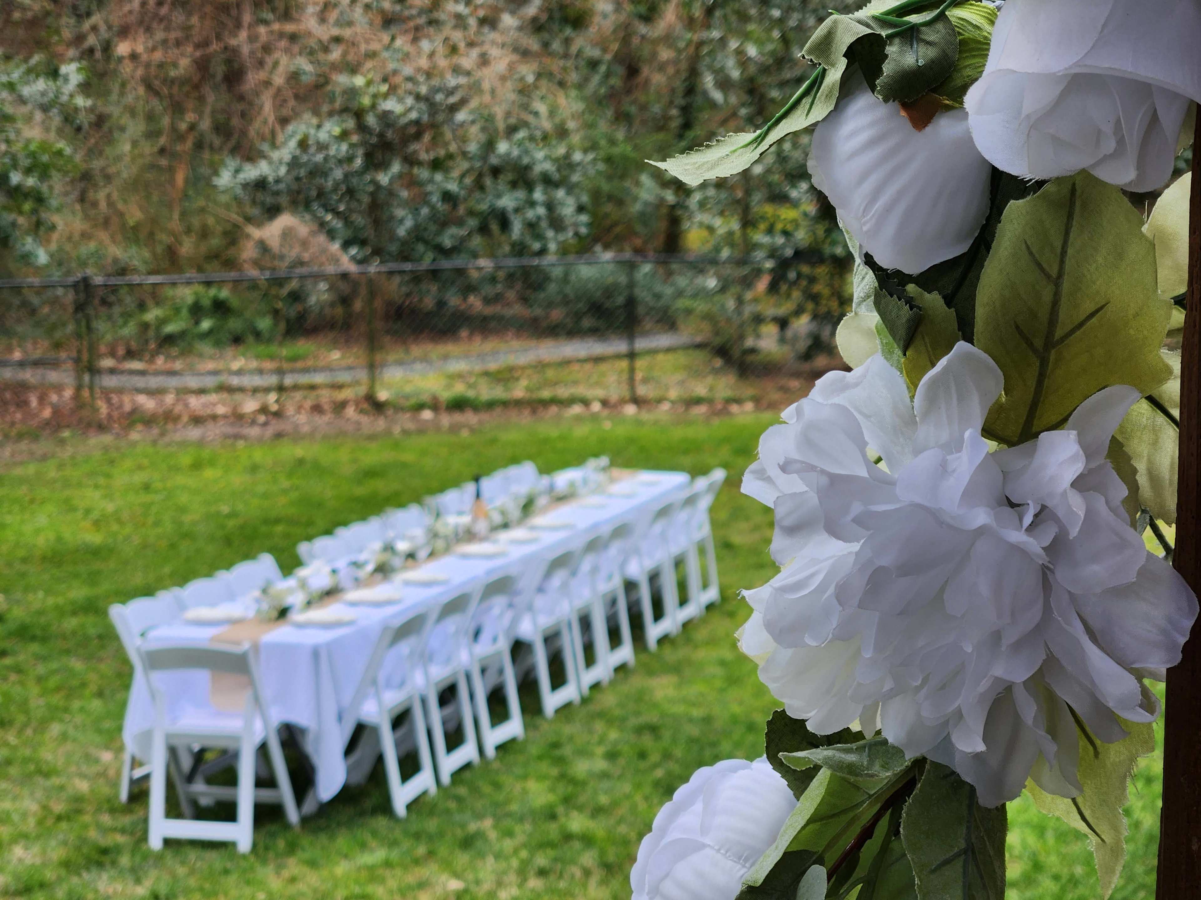 A long table is set with white tablecloths and chairs in a grassy backyard, adorned with white flowers on one side.