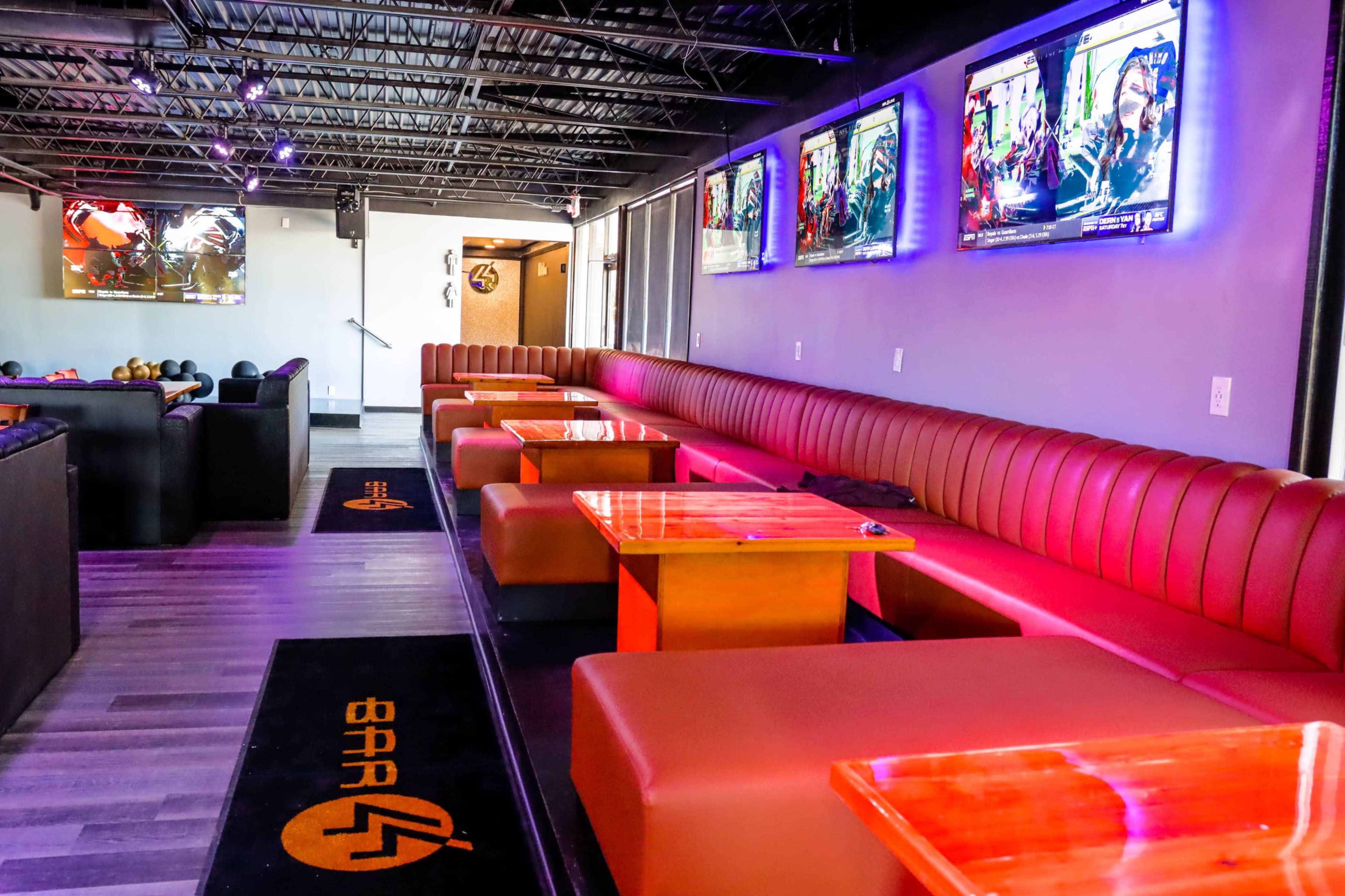 A modern lounge area with plush seating, wooden tables, and multiple screens displaying colorful visuals in a well-lit space.