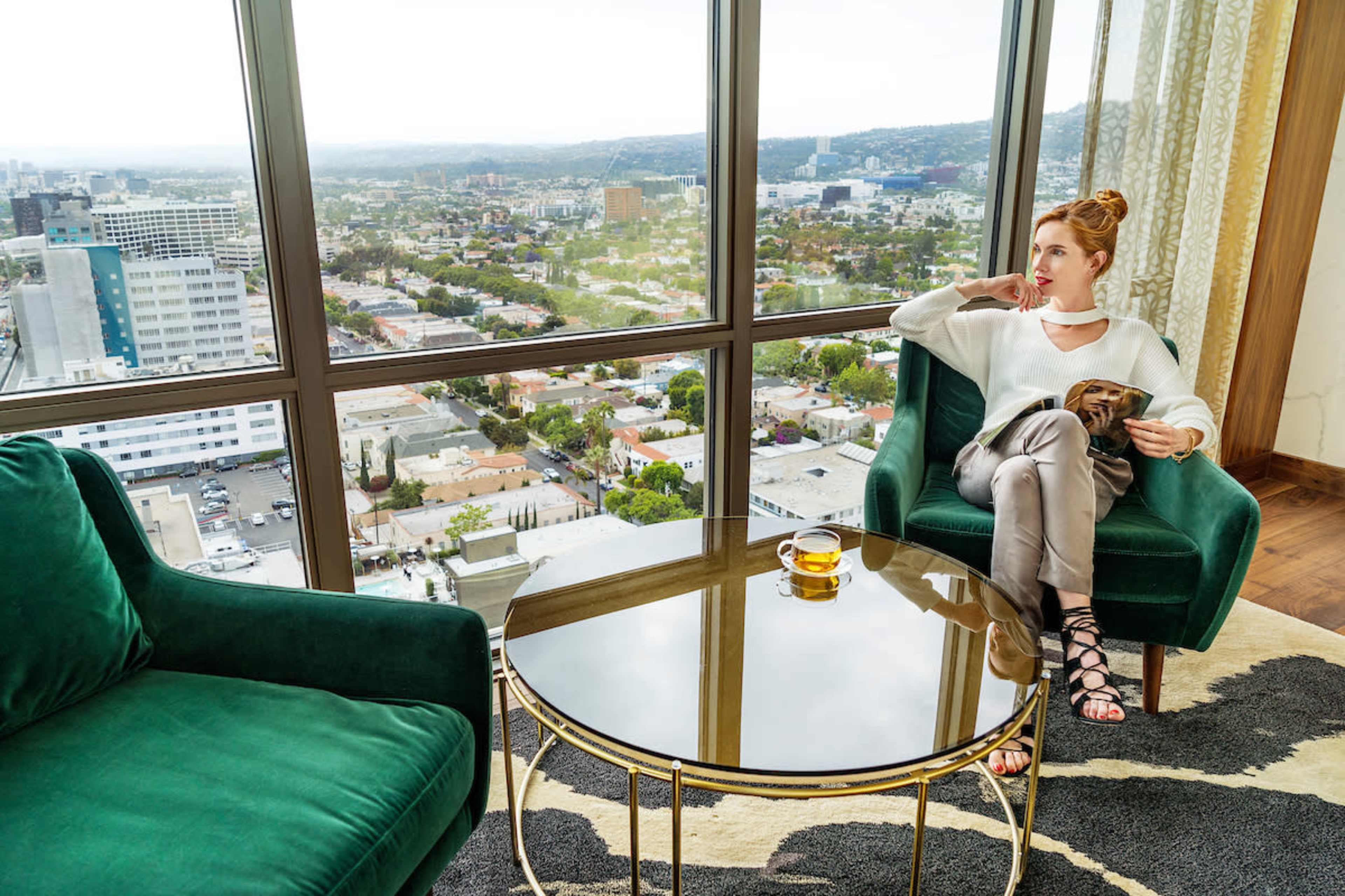 A woman sits in a green armchair near a large window, holding a cup and looking out at the cityscape below.