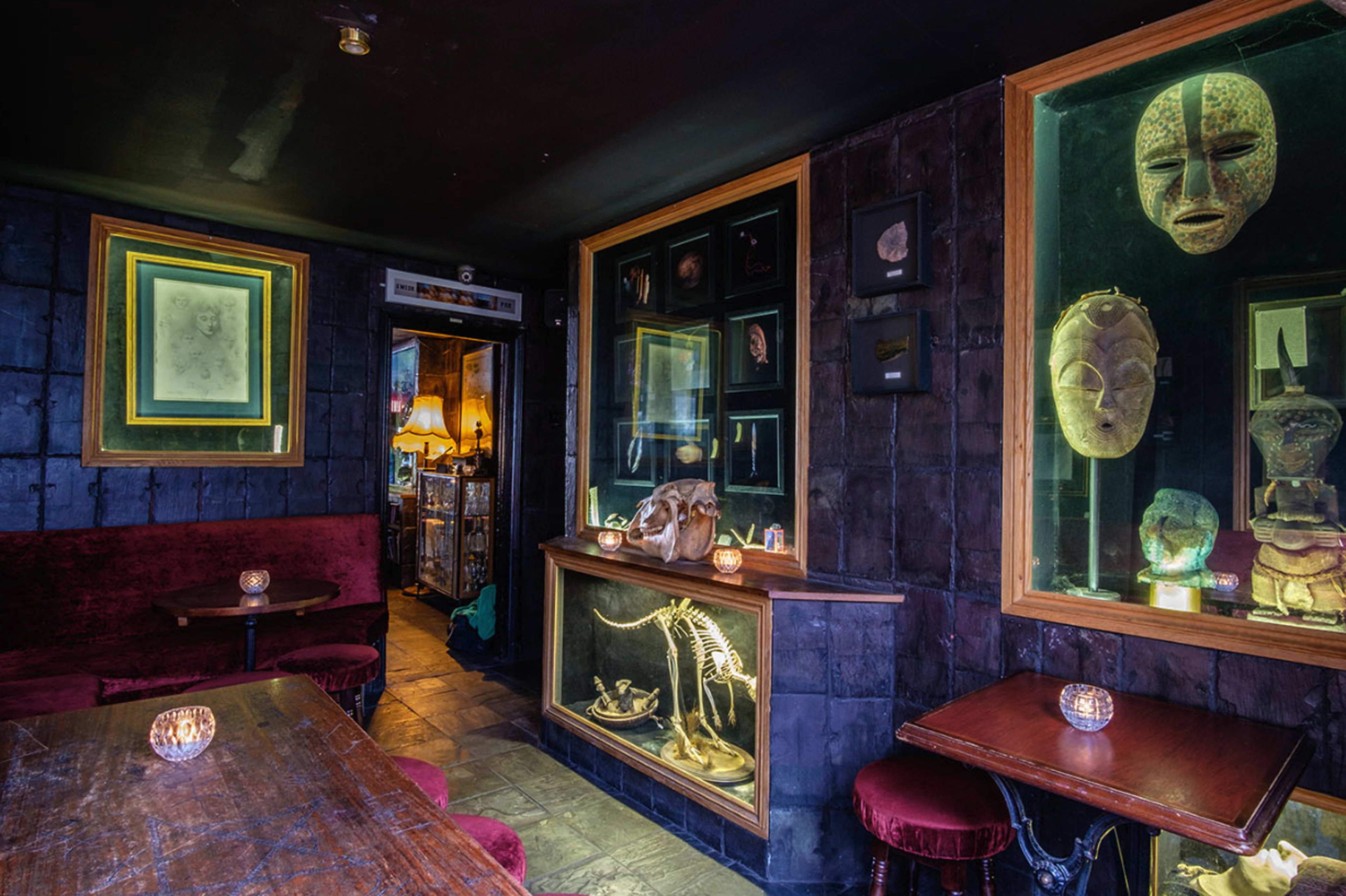 The image depicts a dimly lit bar with dark walls, featuring display cases filled with masks and artifacts, and red velvet seating.