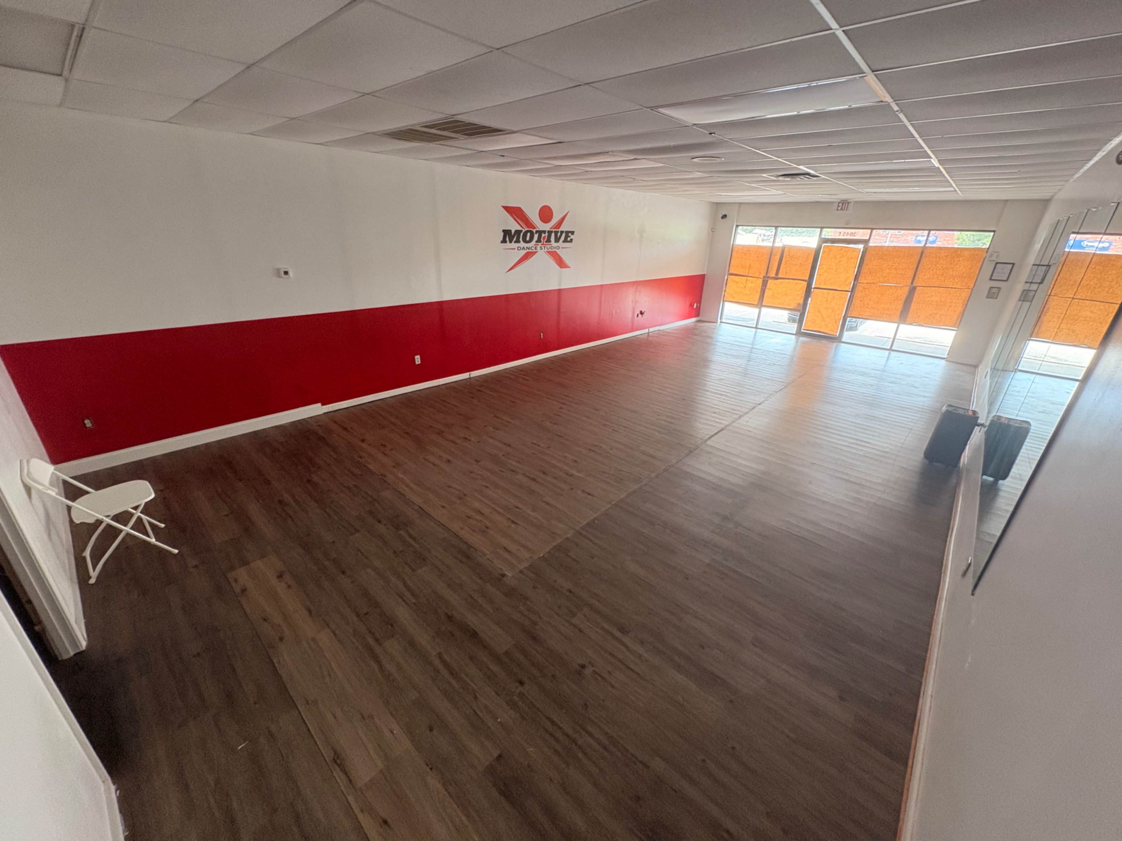 The image shows a spacious, empty interior room with light wood flooring, a red accent wall, and large windows that let in natural light.