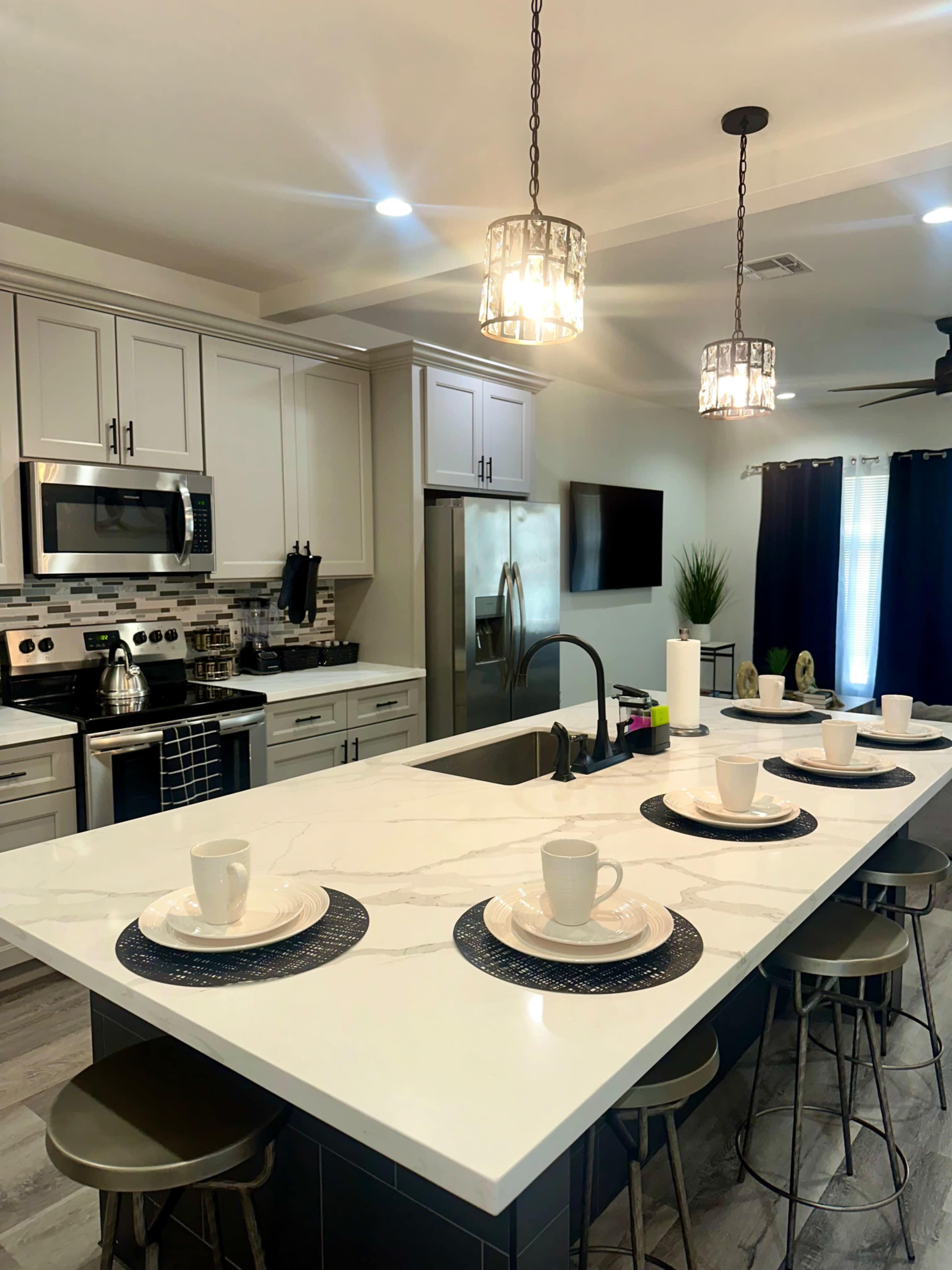 A modern kitchen features an island with place settings and pendant lights, alongside stainless steel appliances and gray cabinetry.