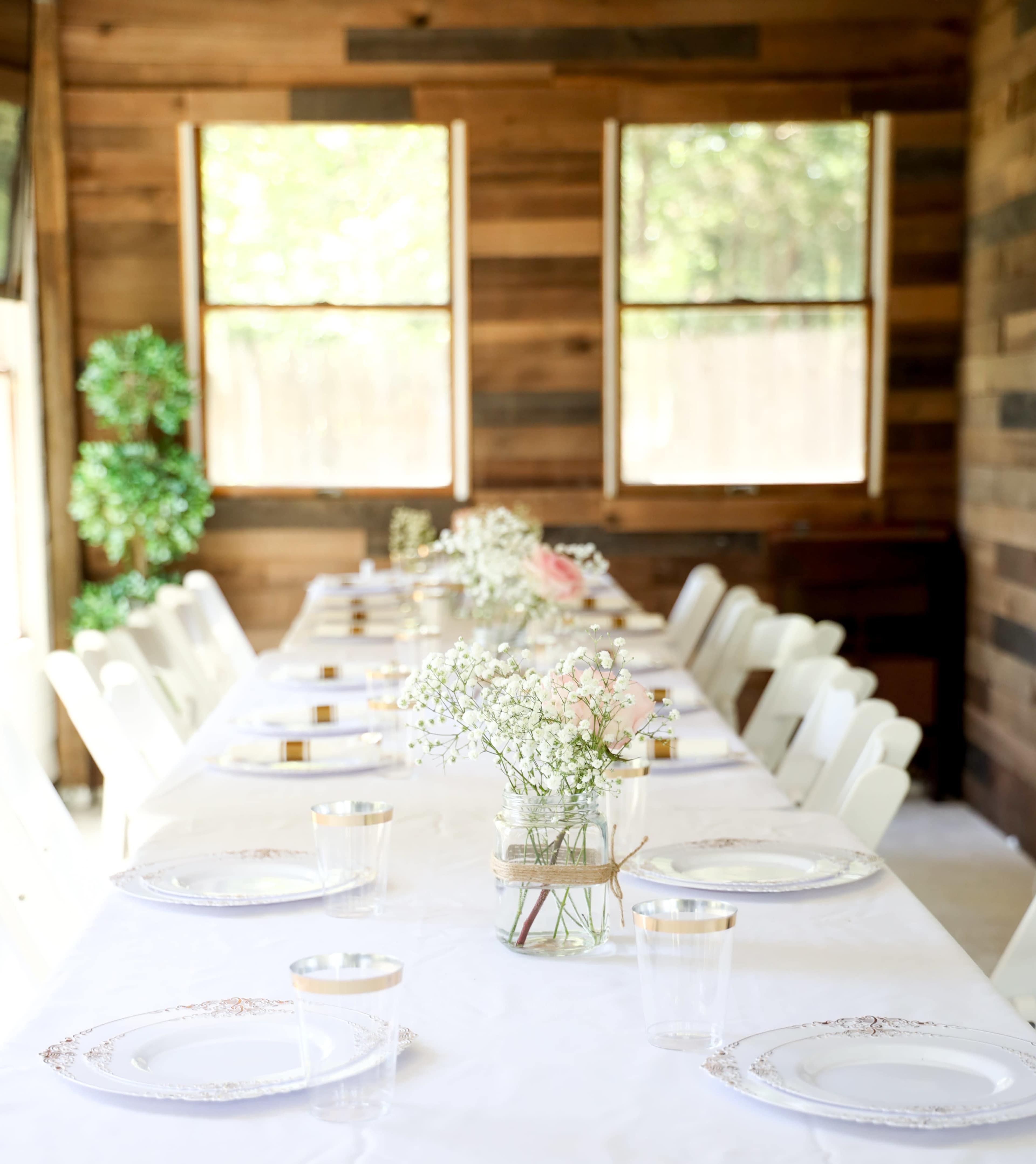A long dining table is set with white plates and flowers in jars, positioned in a room with wooden walls and large windows.