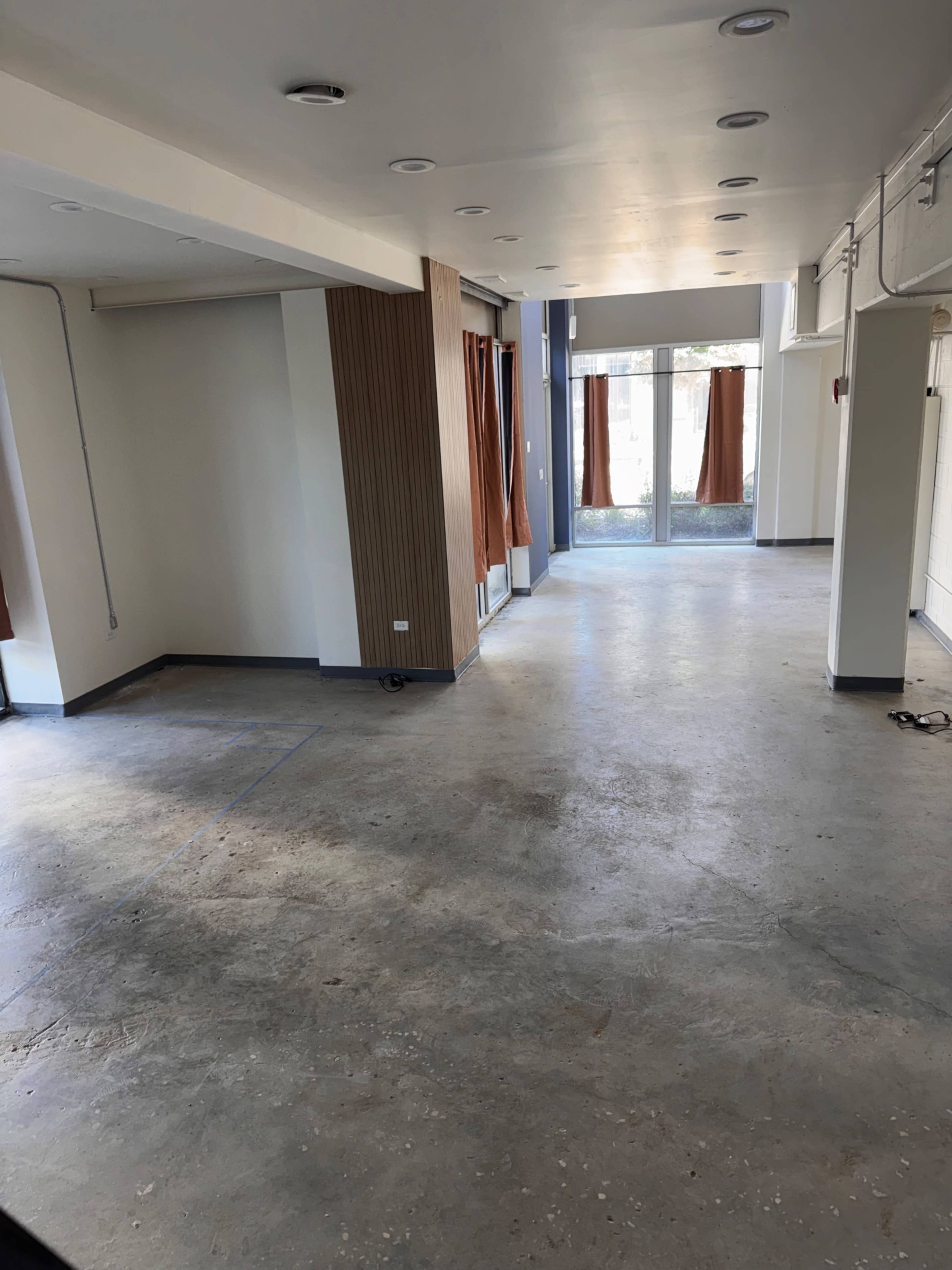 The image shows a spacious interior of a building with concrete flooring, large windows, and minimal furnishings.