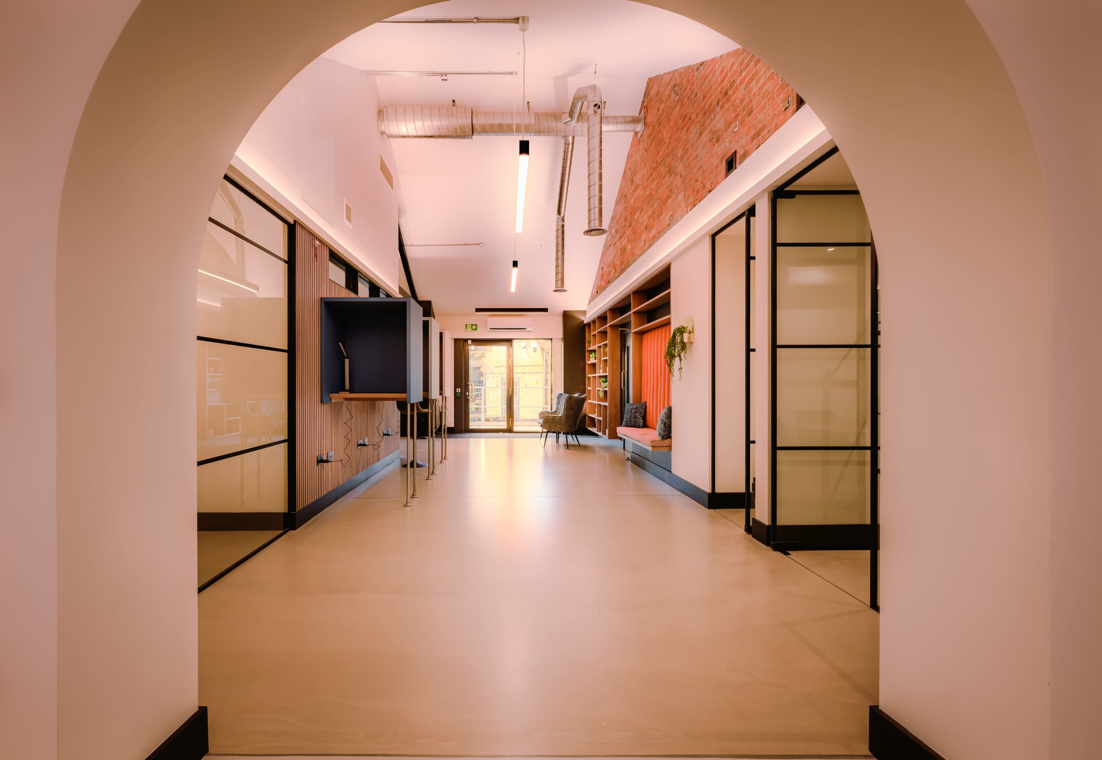 A hallway with large glass doors opens to a bright room featuring a bookshelf and seating area, framed by an archway.