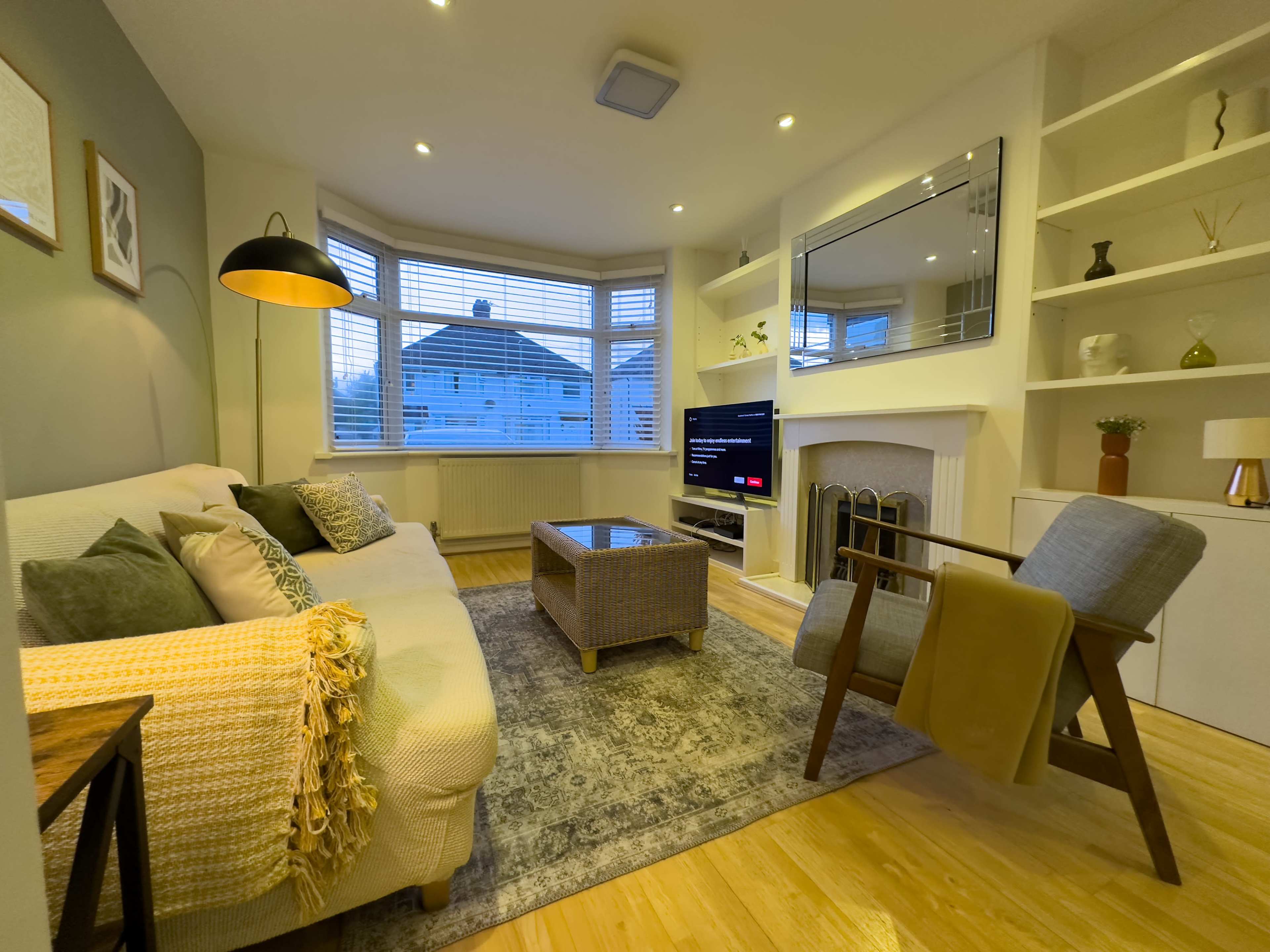 A cozy living room features a light-colored sofa with decorative pillows, a wooden coffee table, and a television mounted on the wall, surrounded by bookshelves.