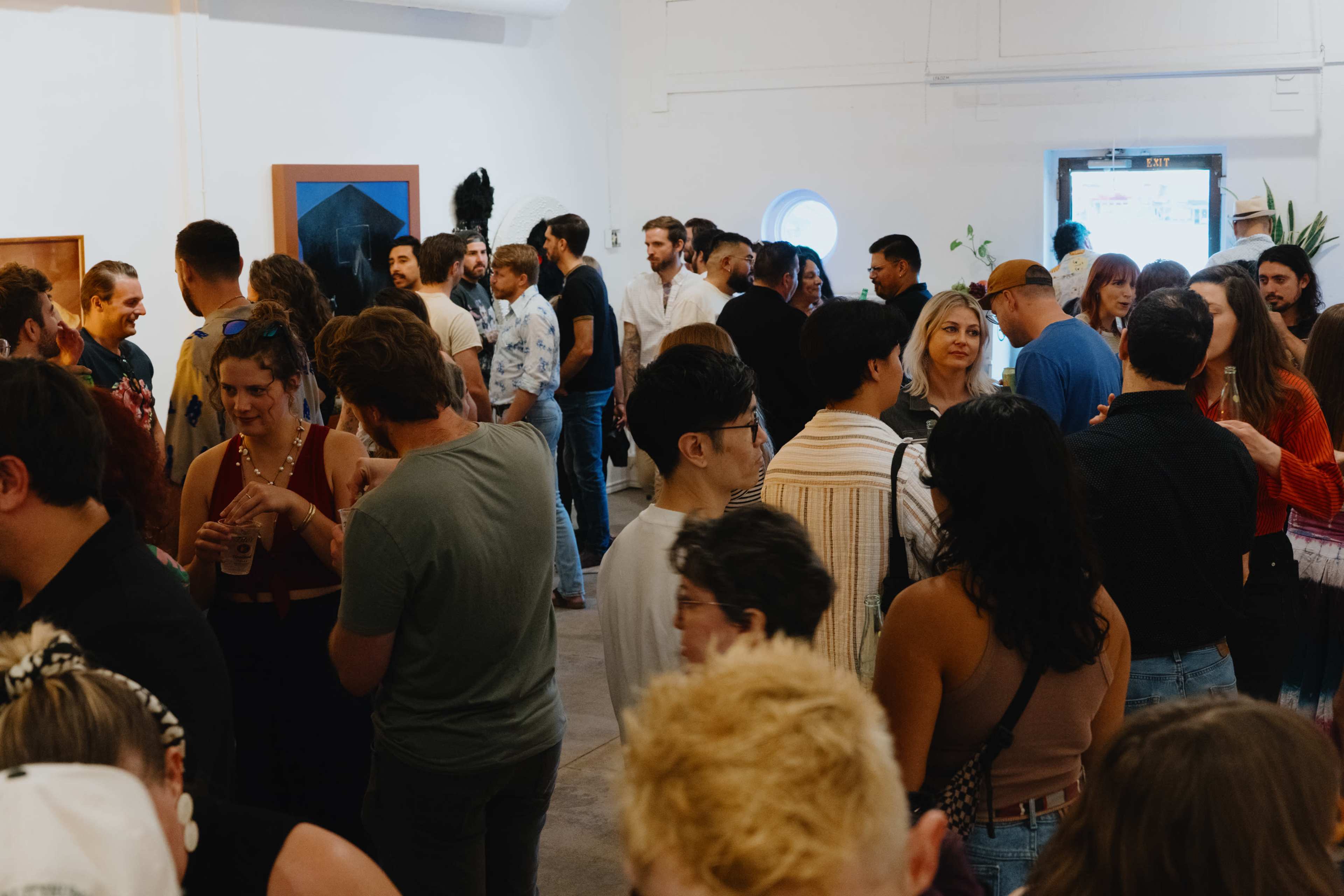 A large group of people is engaged in conversation at an indoor social event, surrounded by art displays and plants.