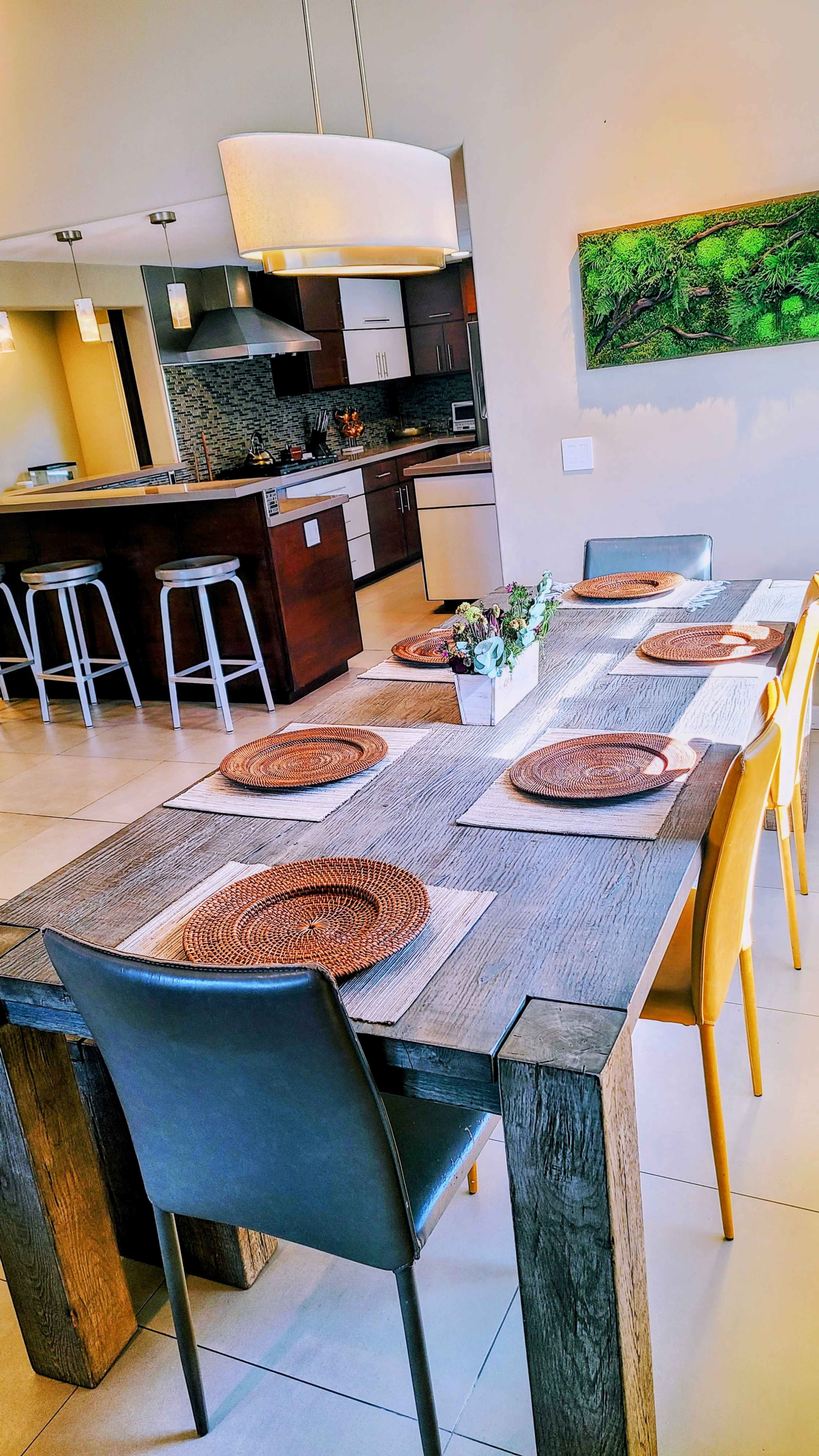 A dining table is set with placemats and surrounded by chairs, with a modern kitchen visible in the background.