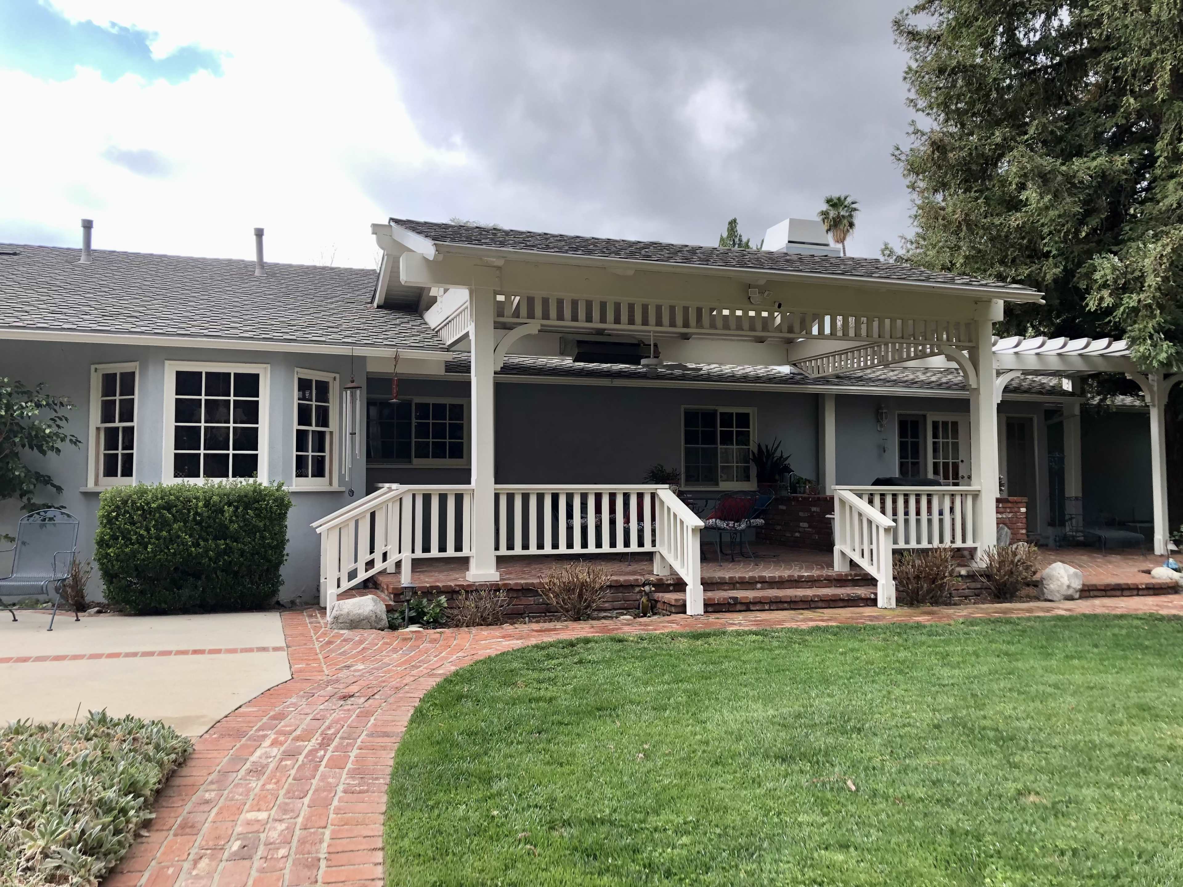 A single-story house with a covered porch and brick walkway is surrounded by green grass and shrubs.