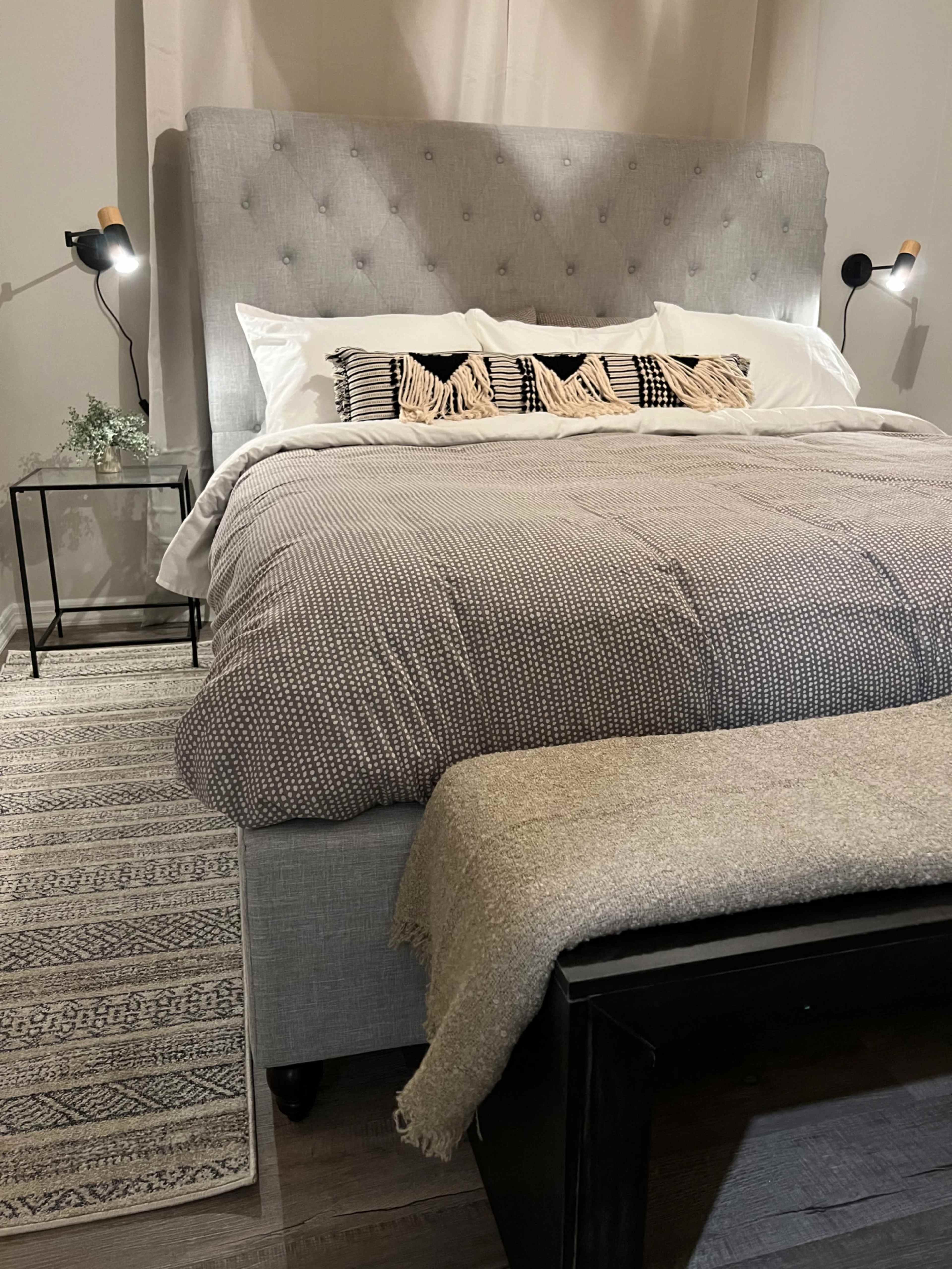 A neatly made bed with a gray upholstered headboard, a patterned blanket, and decorative pillows, accompanied by a small nightstand and a bench at the foot of the bed.