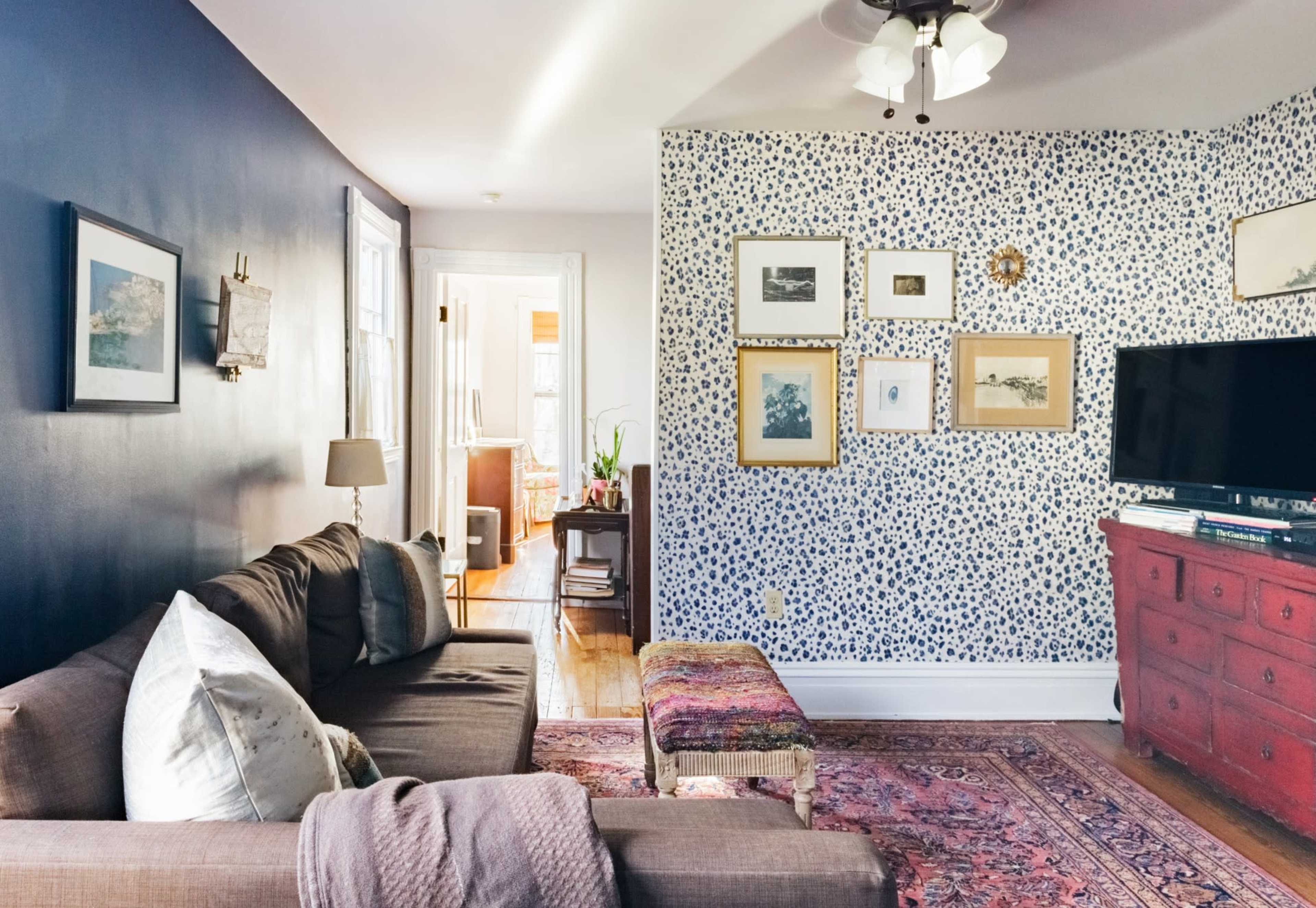 A cozy living room features a dark blue wall and a patterned accent wall, with a brown sofa, a colorful rug, and a television on a red cabinet.