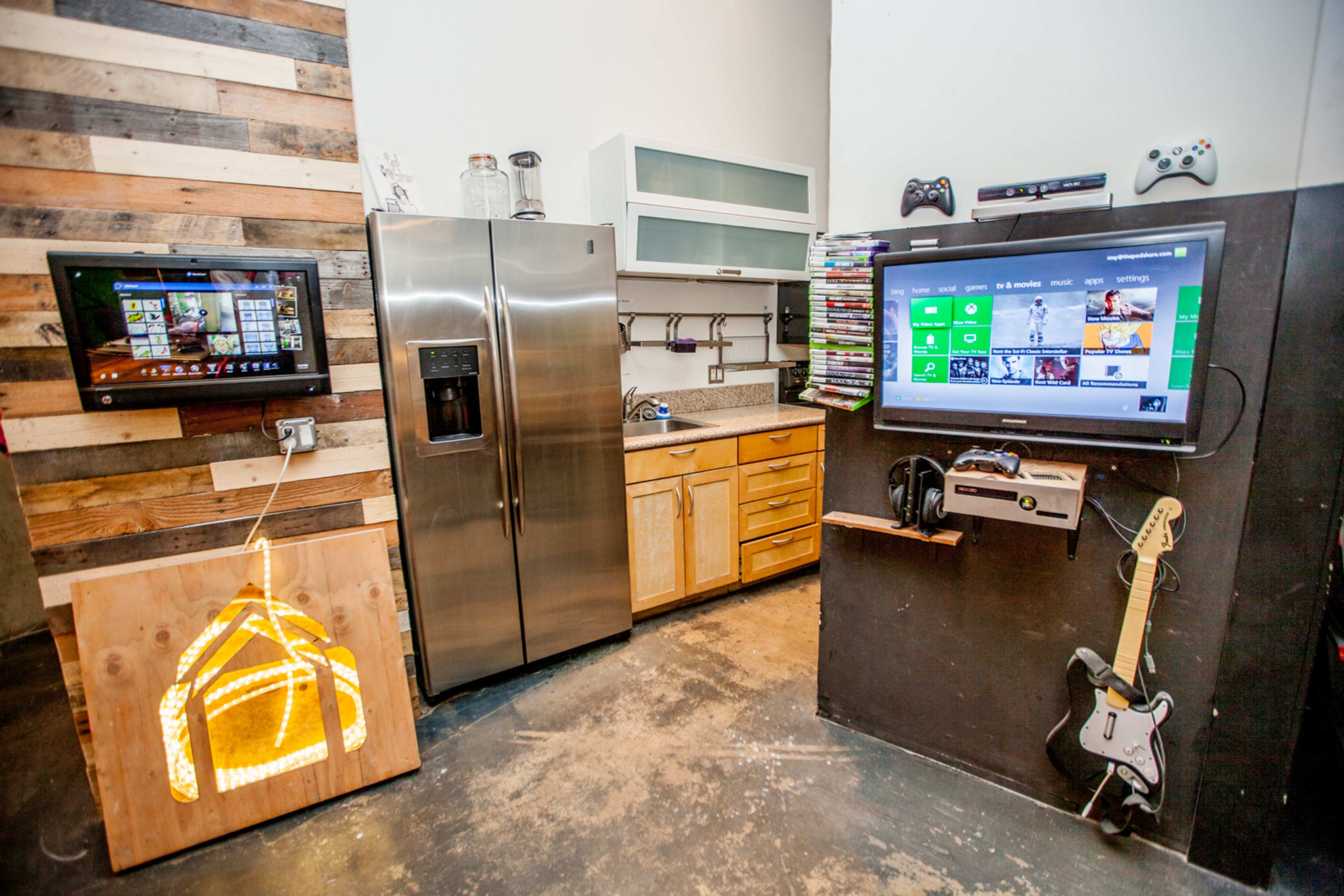 A modern kitchen area featuring a stainless steel refrigerator, wooden cabinets, two wall-mounted televisions, and an illuminated wooden sign, with a guitar leaning against the wall.