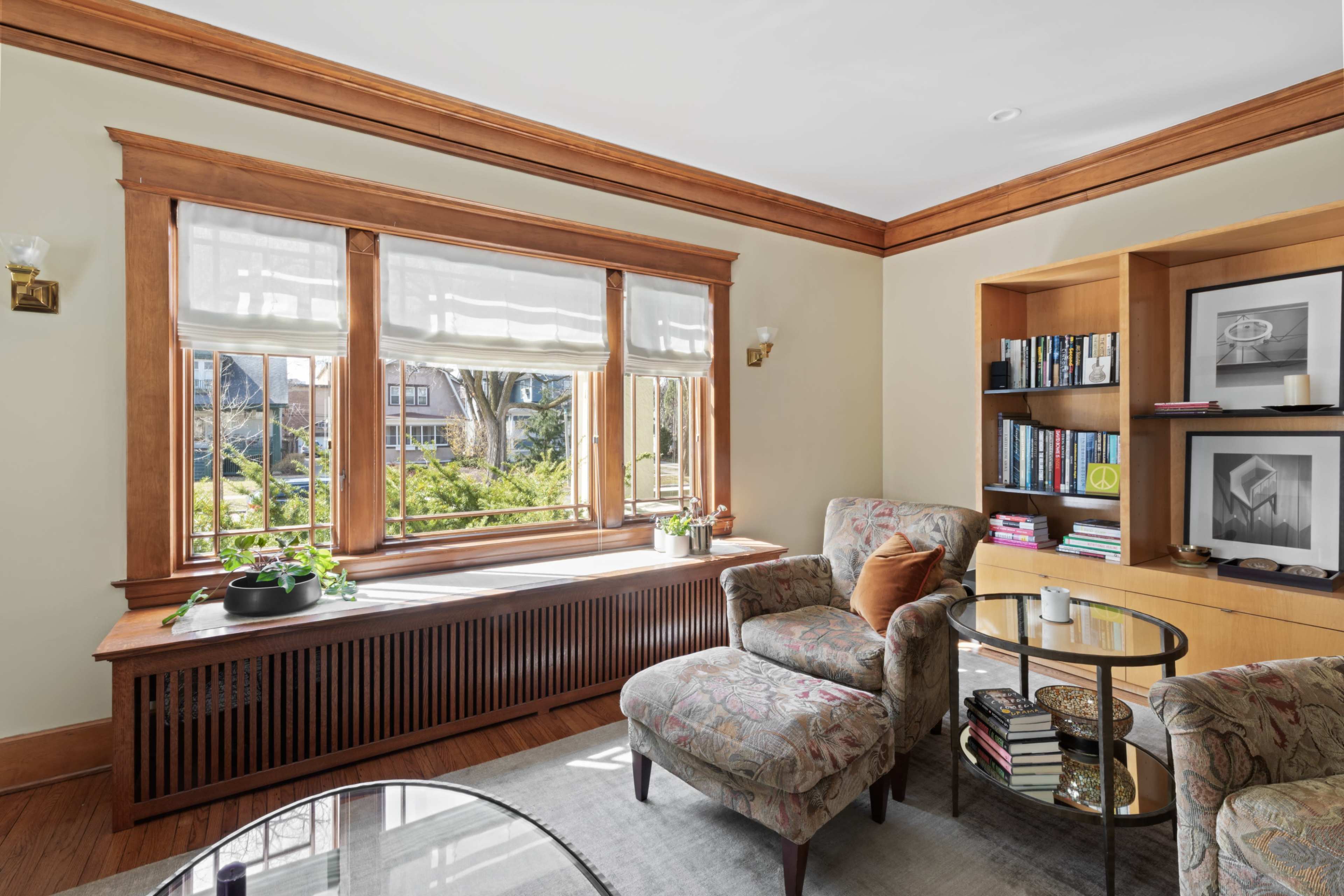 A comfortable living room features a large window with wooden trim, offering a view of greenery outside, and includes upholstered chairs and a bookshelf.