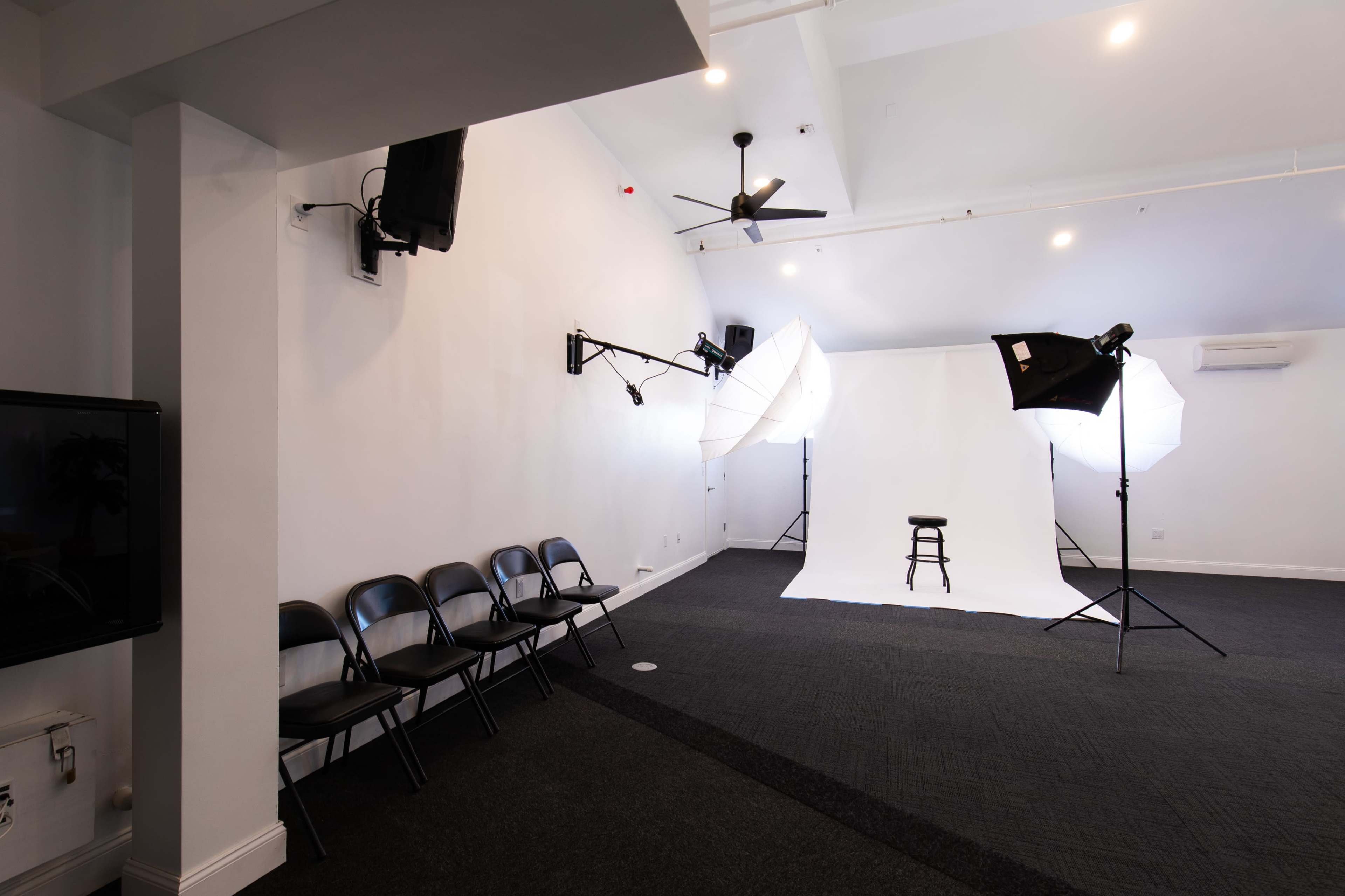 A photography studio with a simple white backdrop, lighting equipment, and rows of black chairs along the side.