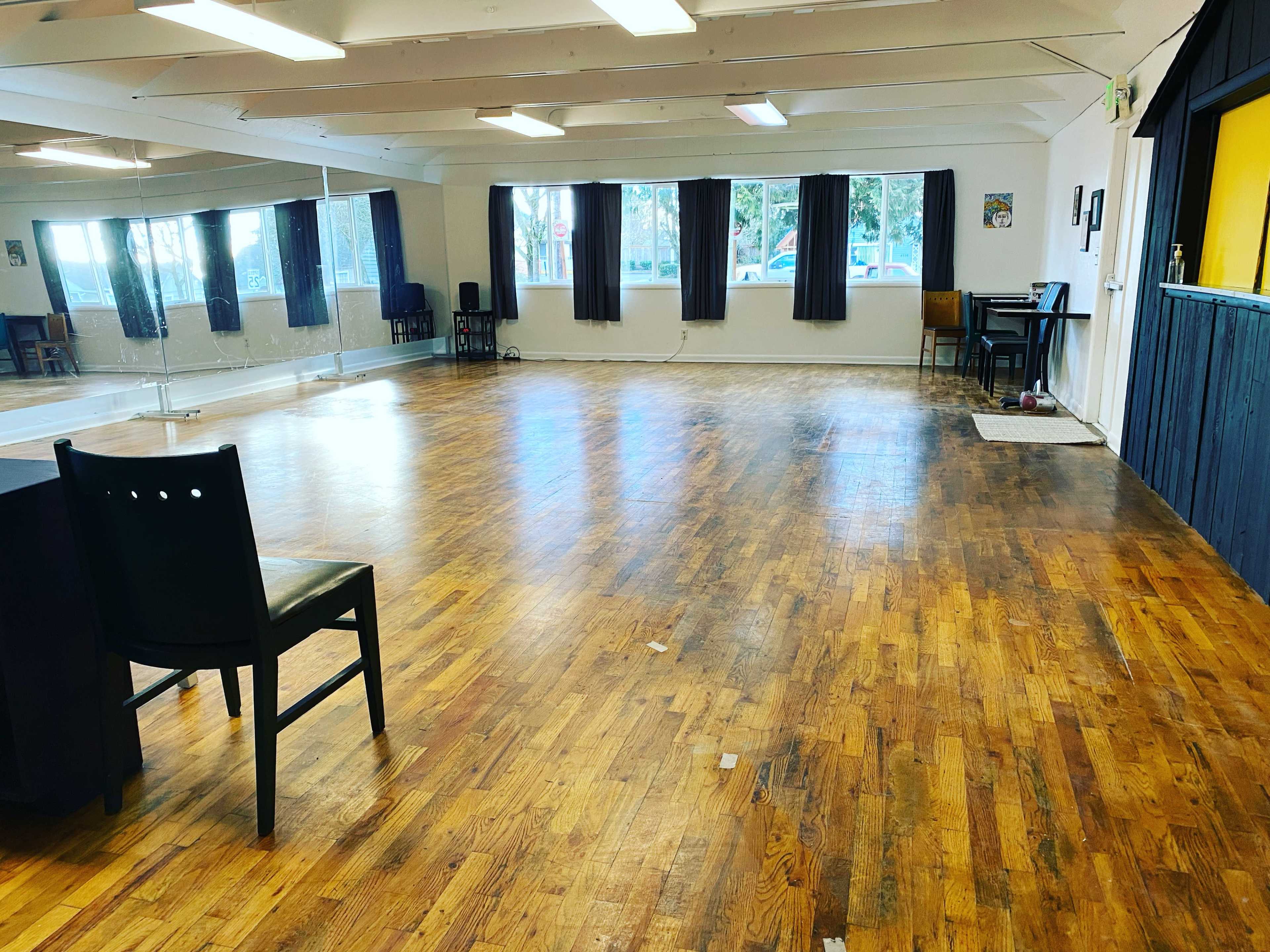 A spacious dance studio with wooden floors, mirrors on one wall, and several windows allowing natural light.
