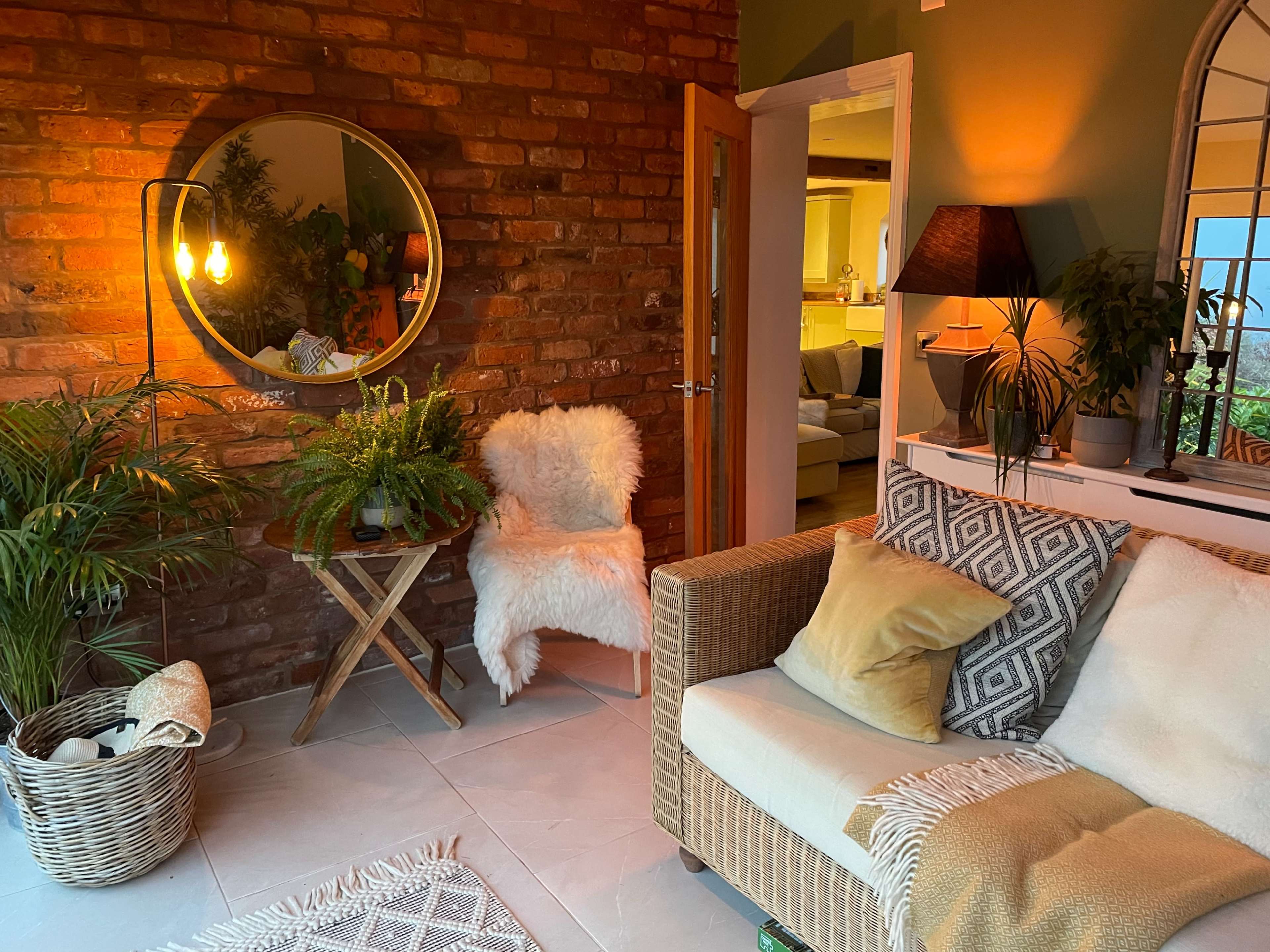 A cozy sitting area features a brick wall, a mirror, a soft chair, and a patterned sofa surrounded by indoor plants.