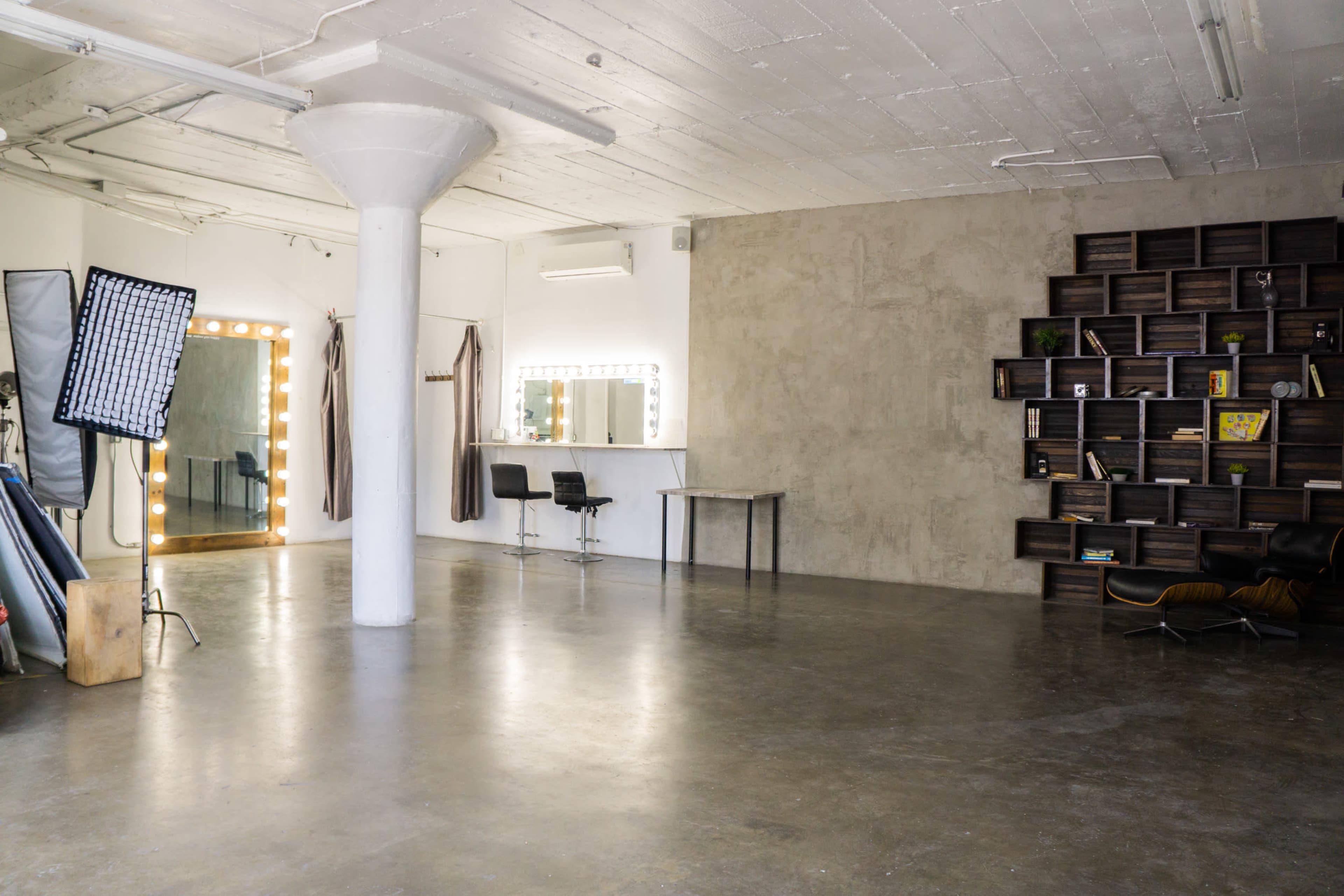 The image shows a spacious, minimalist studio with a concrete floor, a large pillar in the center, a lighted mirror setup, and a wall of wooden shelves filled with various items.