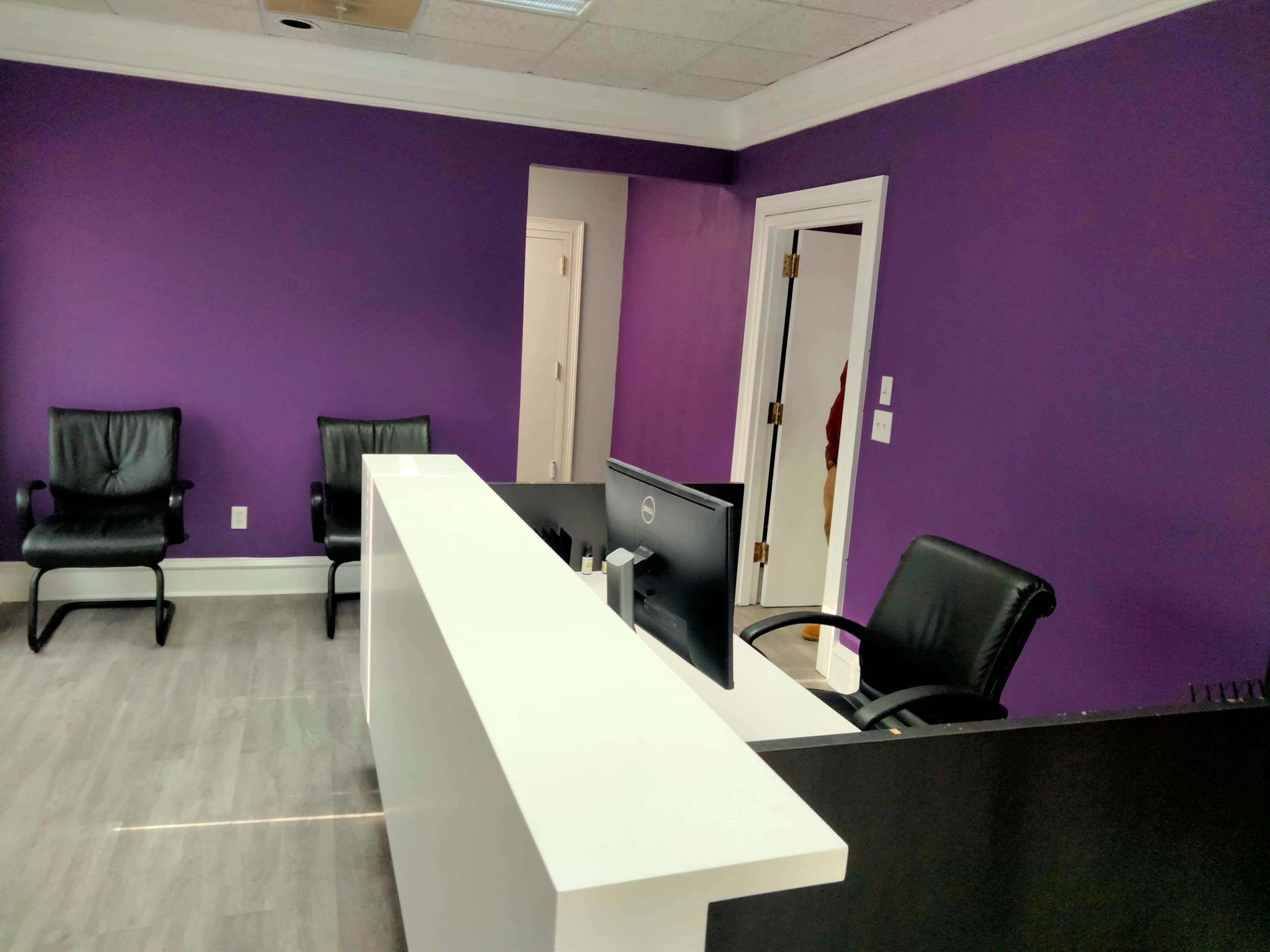The image shows a modern office reception area with a white front desk, a computer monitor, and two black chairs against a purple wall.