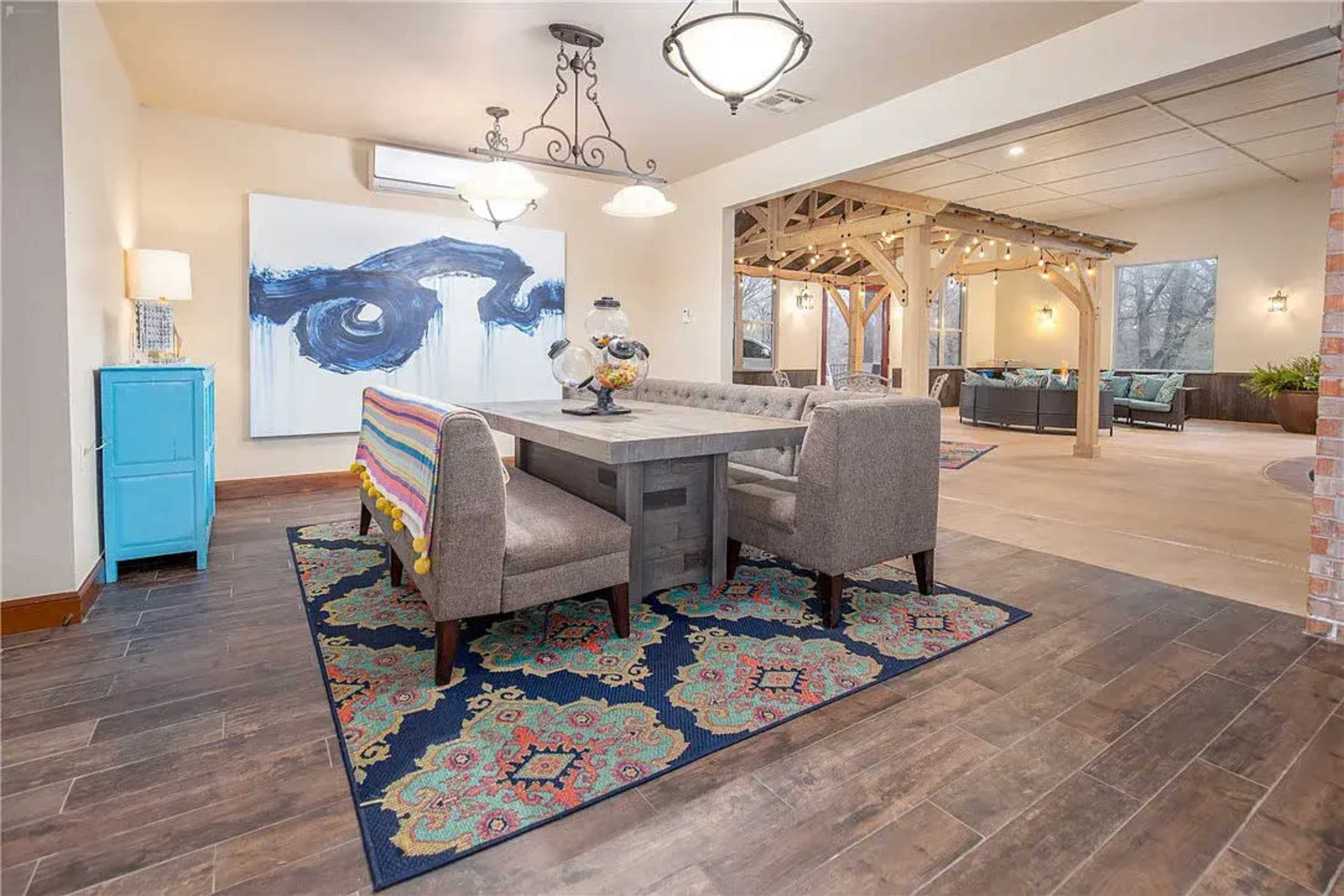 A dining area featuring a rectangular table surrounded by upholstered chairs, with colorful artwork on the wall and a patterned rug beneath the table.