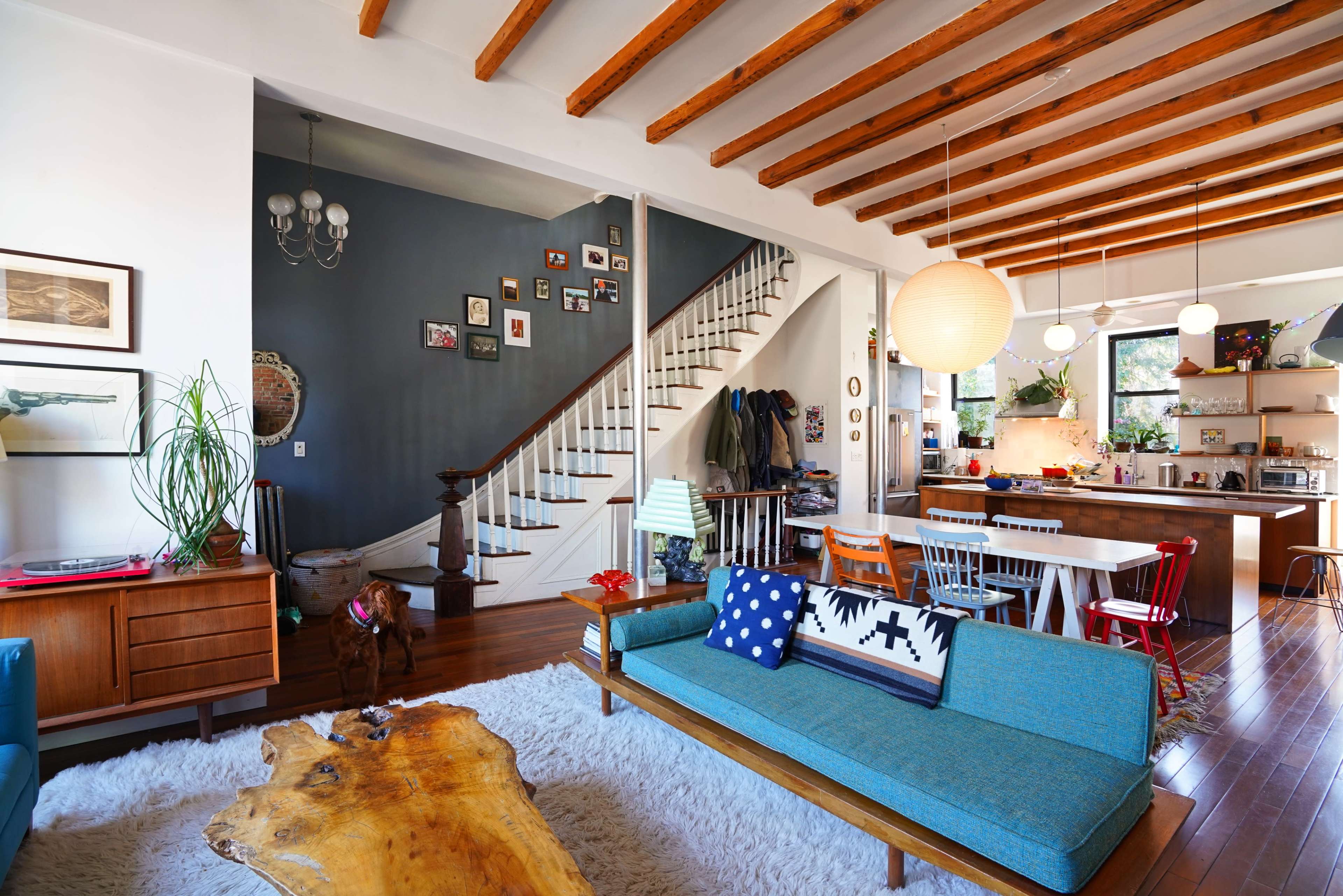 A bright living space with wooden beams, a blue sofa, and an open layout that connects to a staircase and kitchen area.