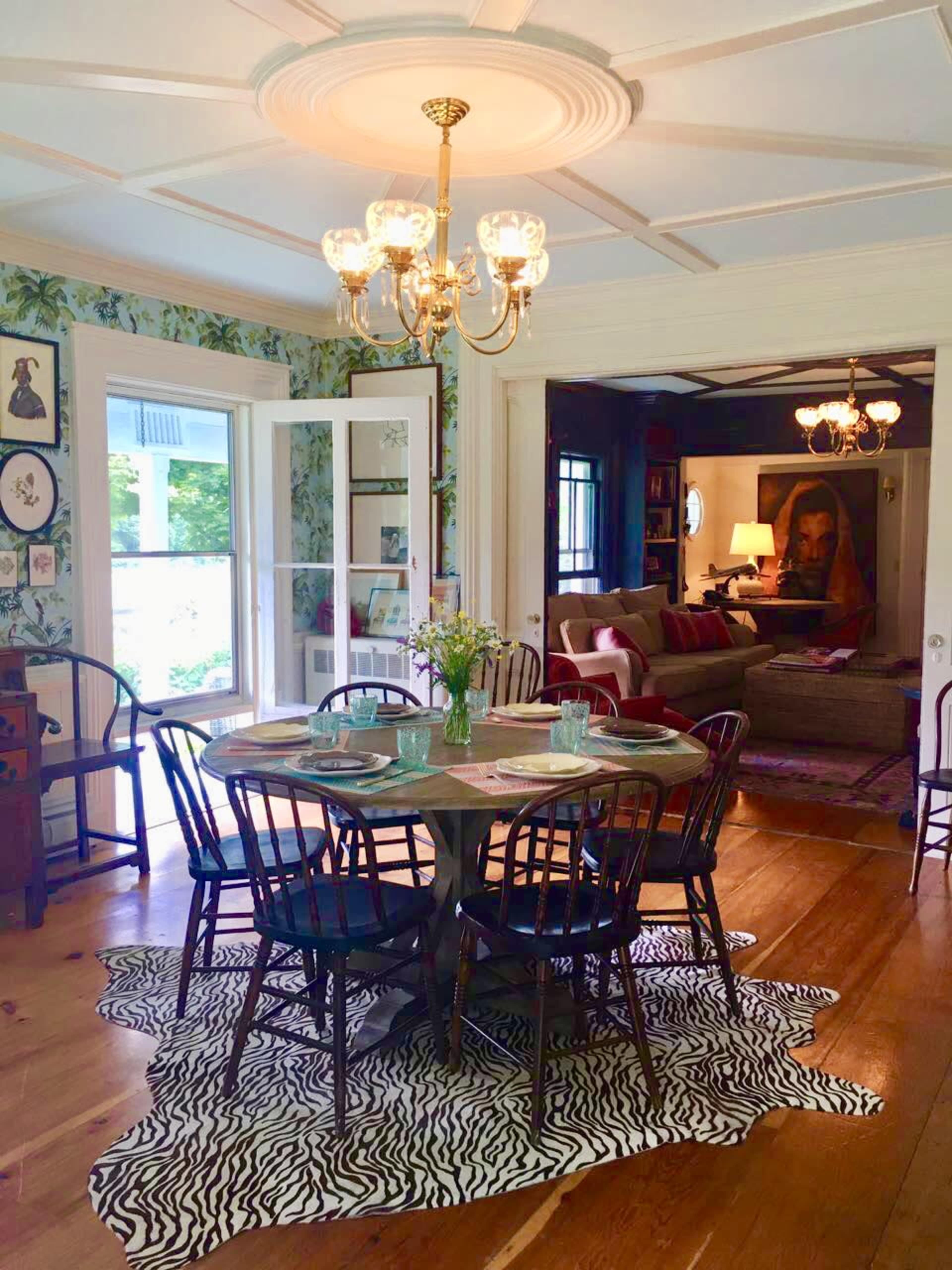A dining area with a round wooden table surrounded by black chairs on a zebra-print rug, with a chandelier above and greenery-patterned wallpaper on the walls.