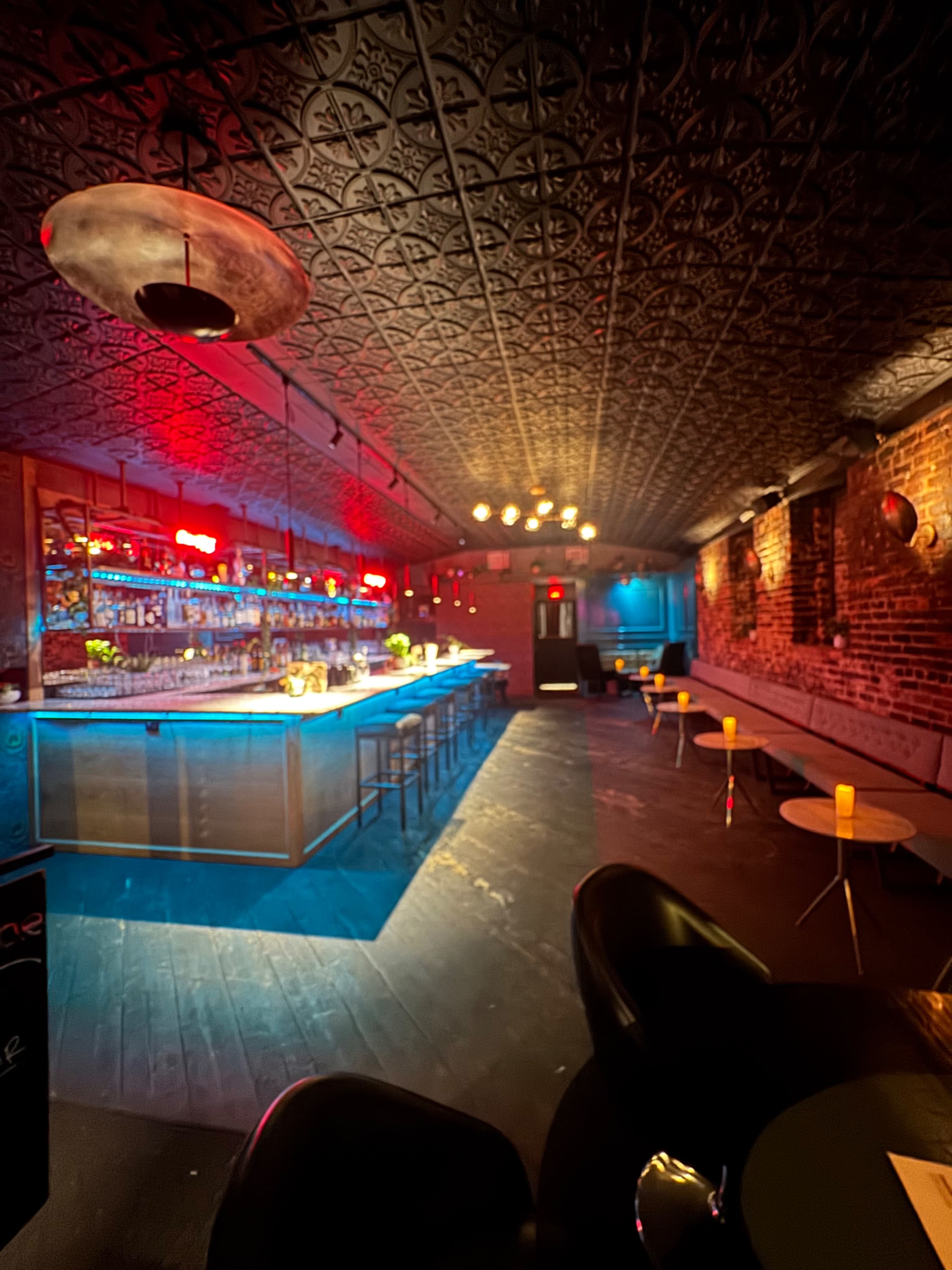 The image shows a dimly lit bar interior with a long illuminated counter, high stools, and exposed brick walls.