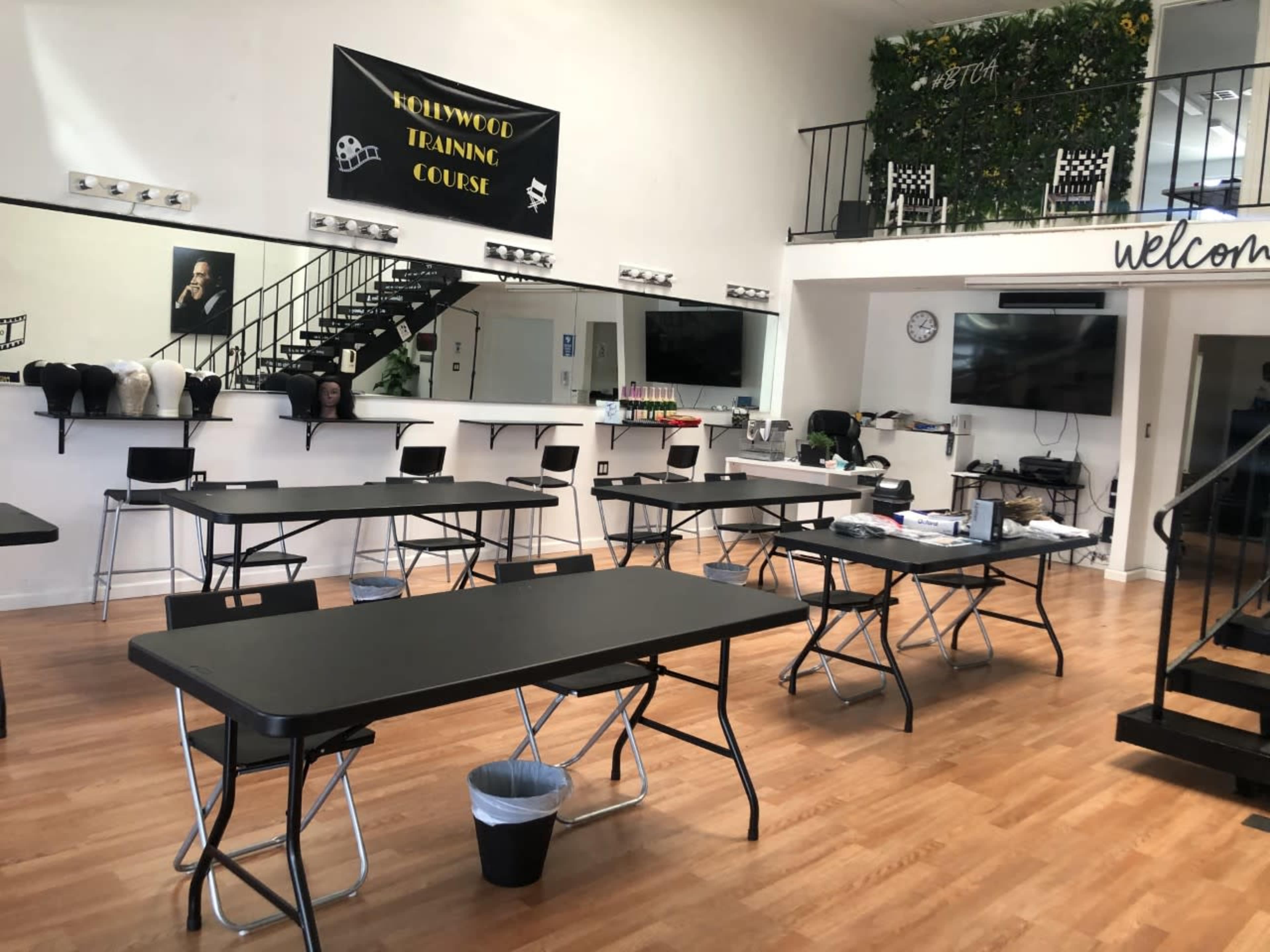 An interior space with several black folding tables arranged in a classroom setup, featuring a banner that reads "Hollywood Training Course" and a green wall in the background.