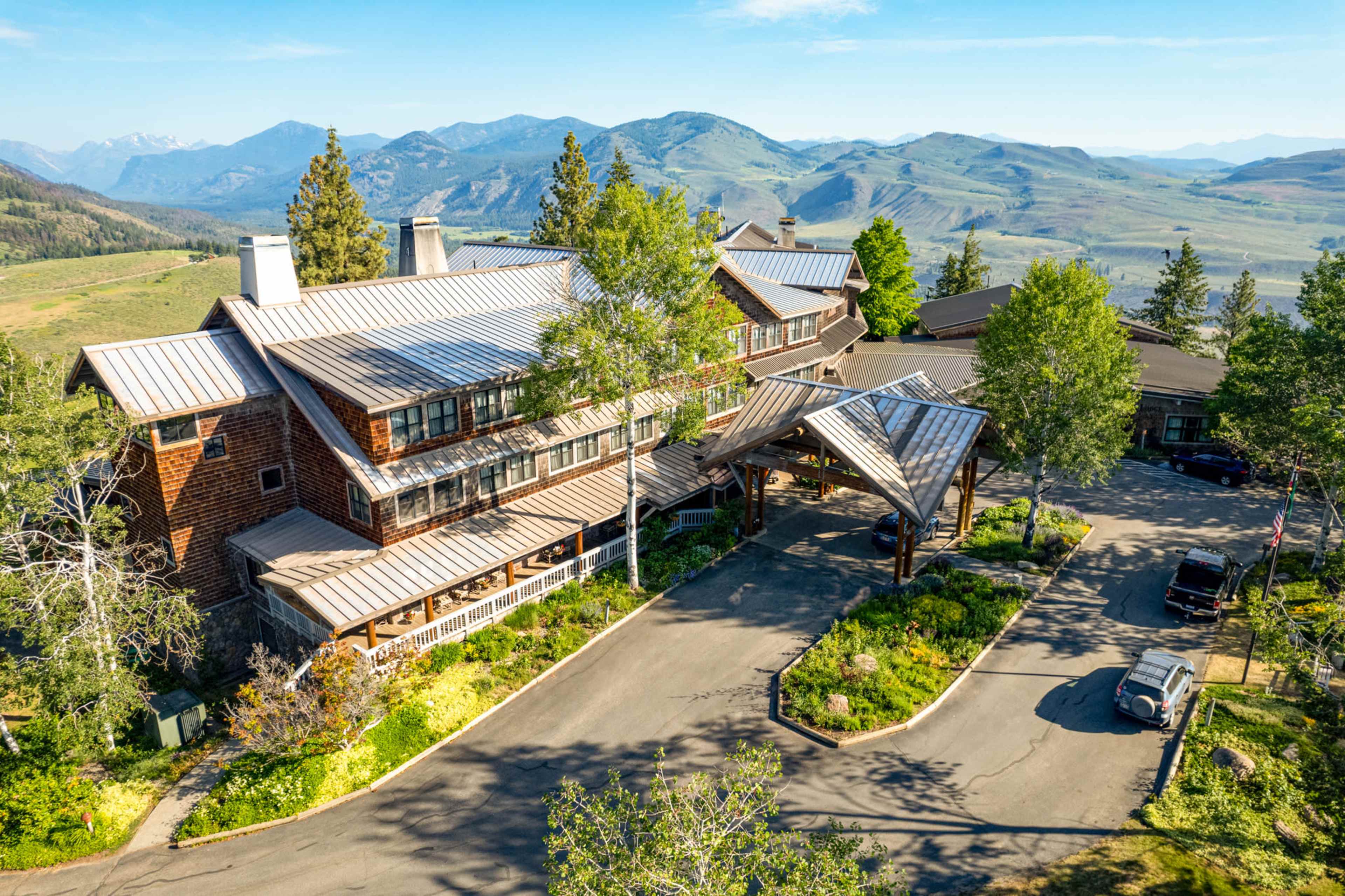 The image shows a large, multi-story lodge surrounded by trees and mountains, with a paved driveway leading up to the entrance.