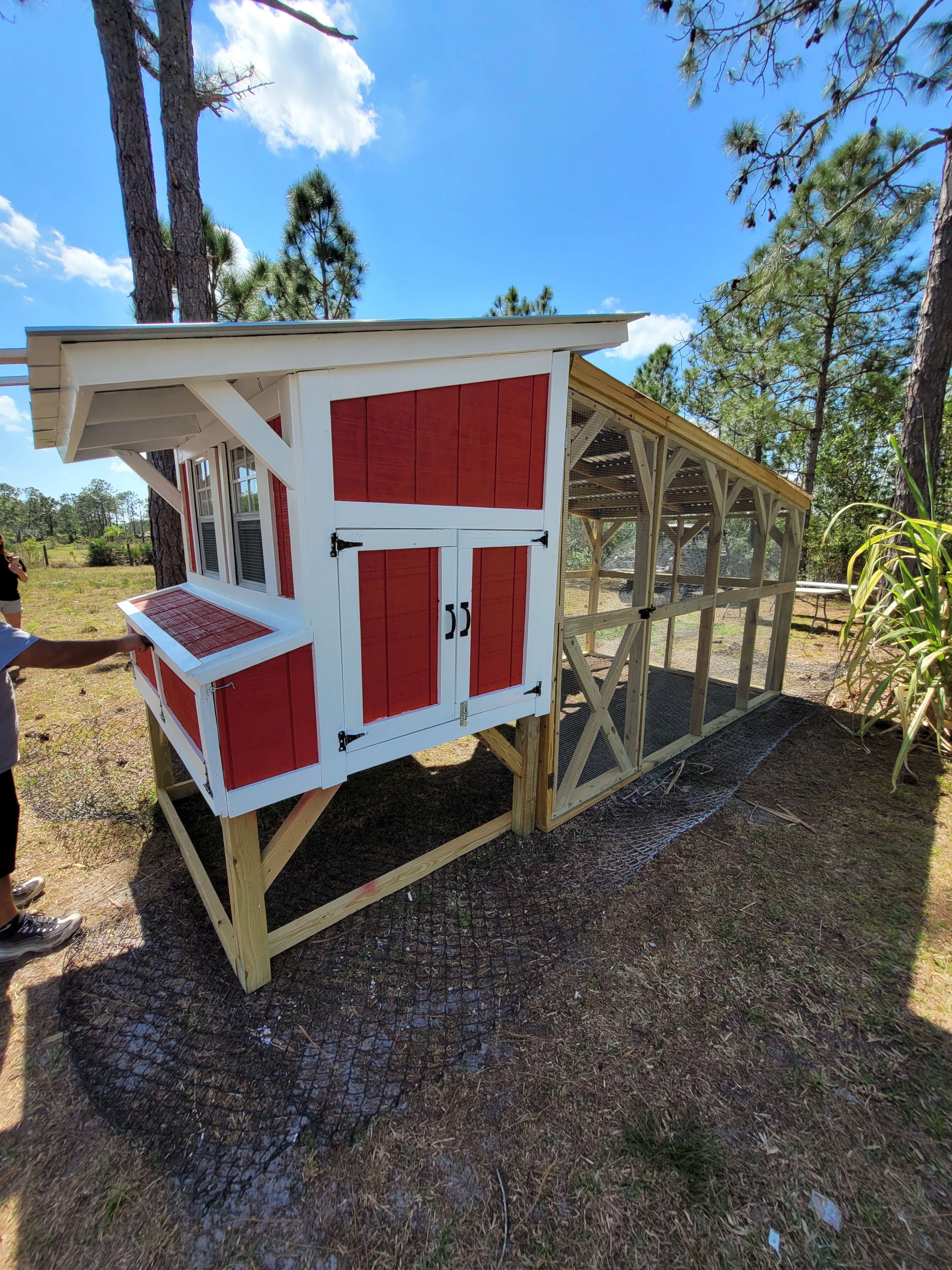 A red and white chicken coop with a connected outdoor run, situated in a grassy area surrounded by trees.