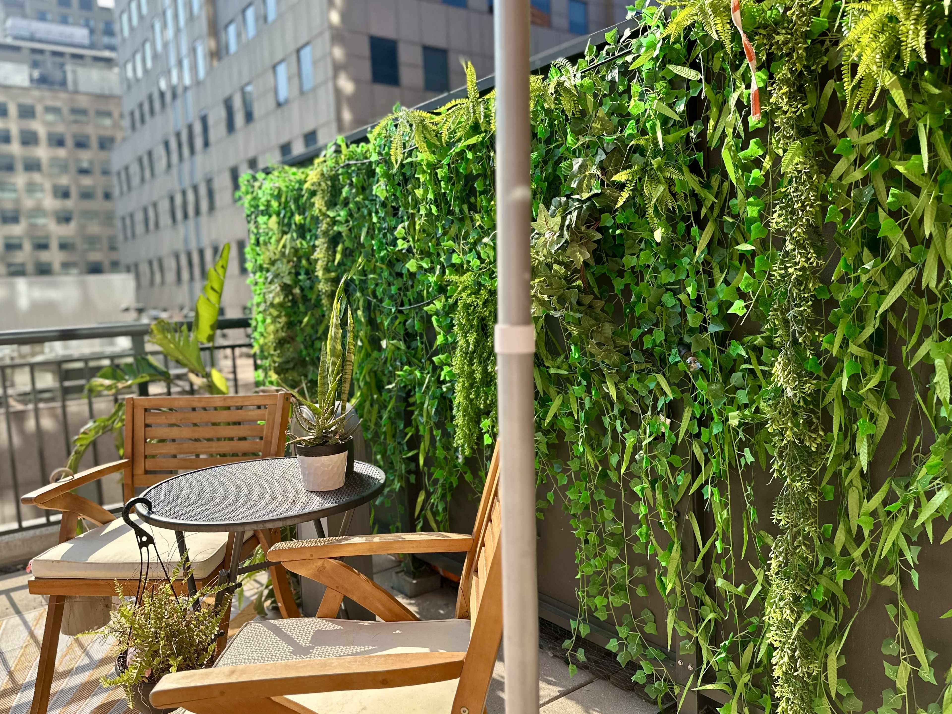 A balcony features wooden chairs and a table surrounded by lush green foliage.