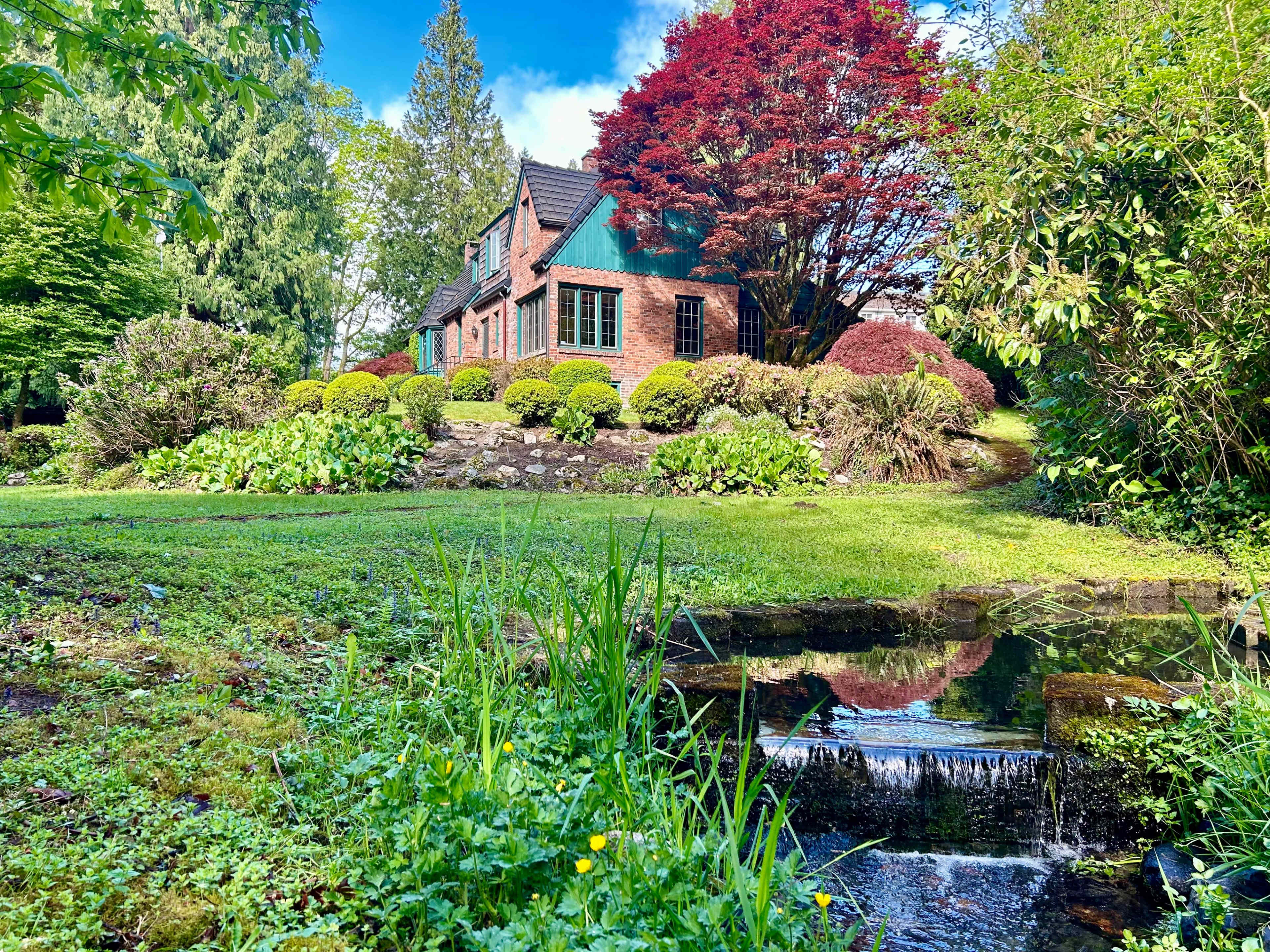 A brick house with green accents is surrounded by manicured gardens and a small stream flows nearby.
