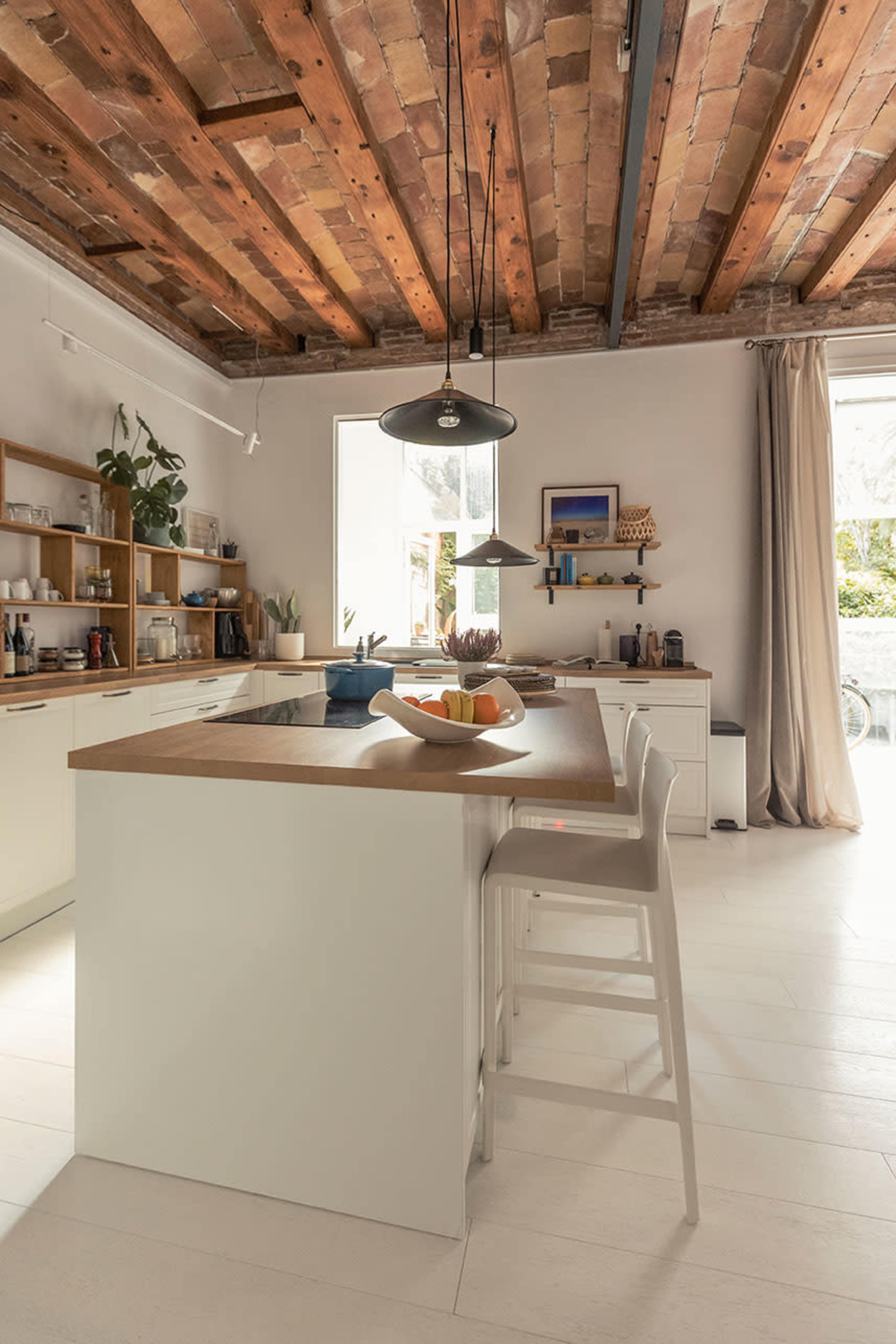A modern kitchen features an island with seating, wooden beams overhead, and ample natural light streaming through a large window.