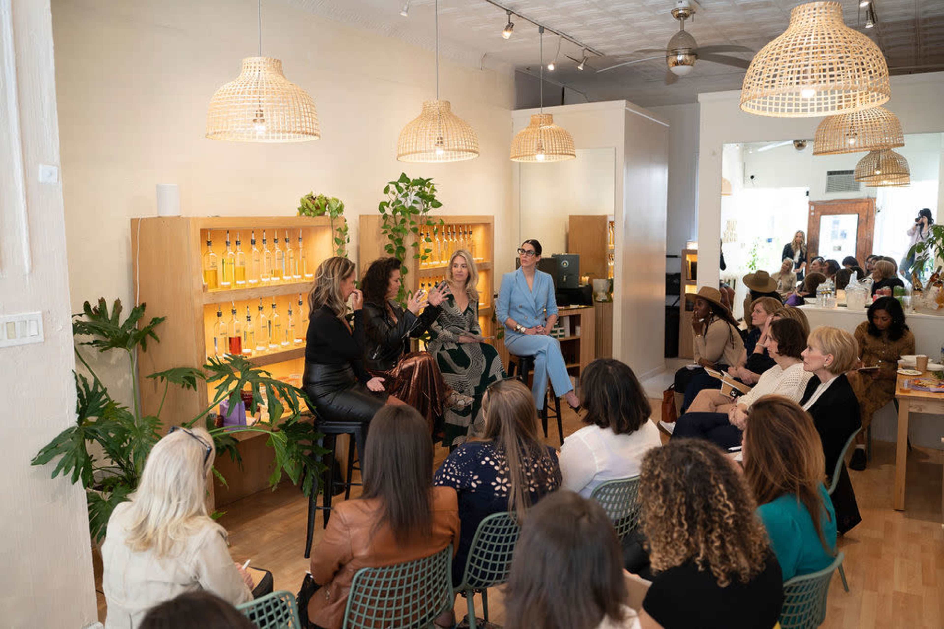 A group of women participates in a panel discussion in a bright, airy room filled with plants and shelves of products.